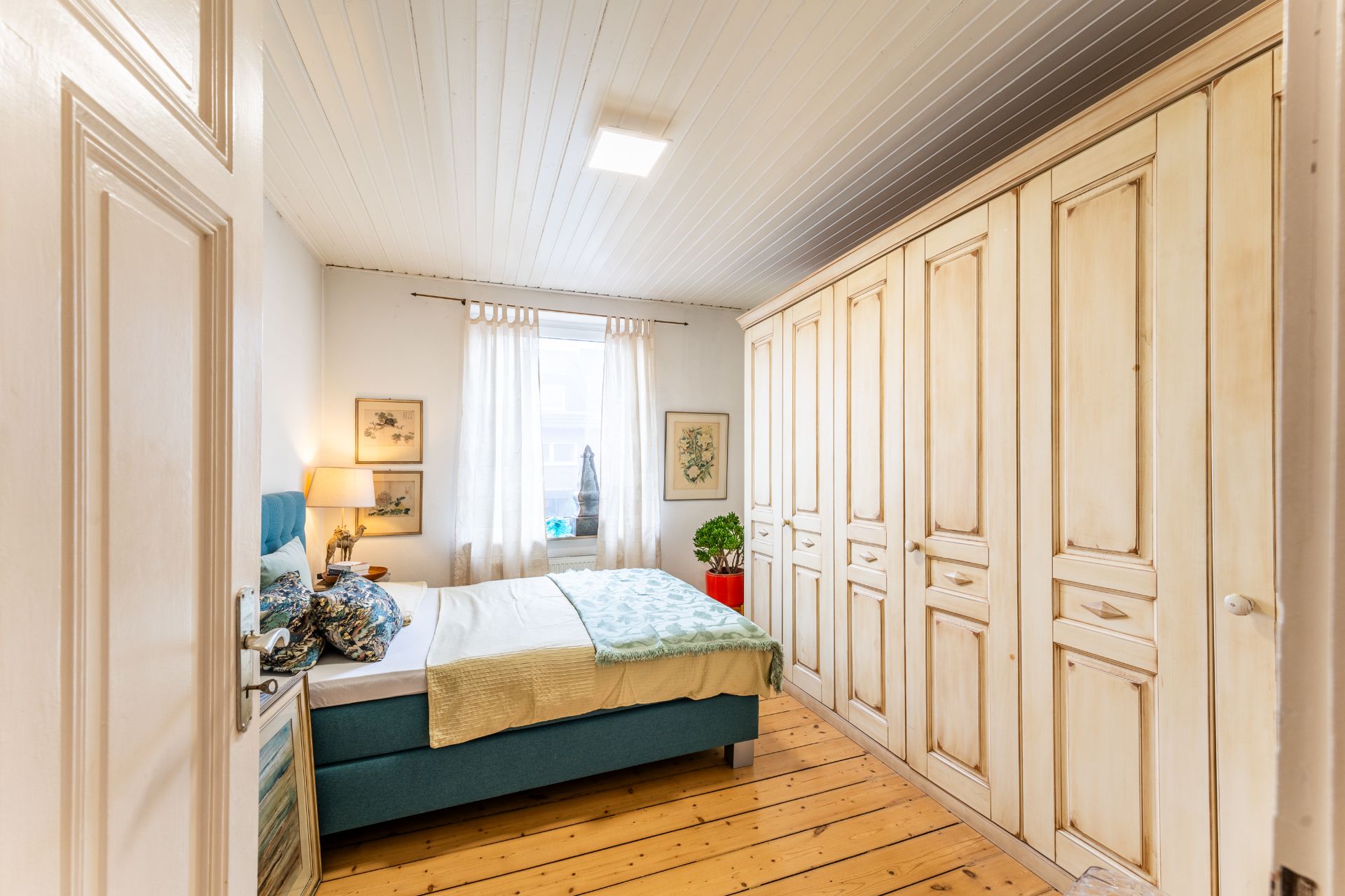 Helles Schlafzimmer mit großem Holzschrank, Bett, Nachttisch und Fenster mit weißen Vorhängen.
