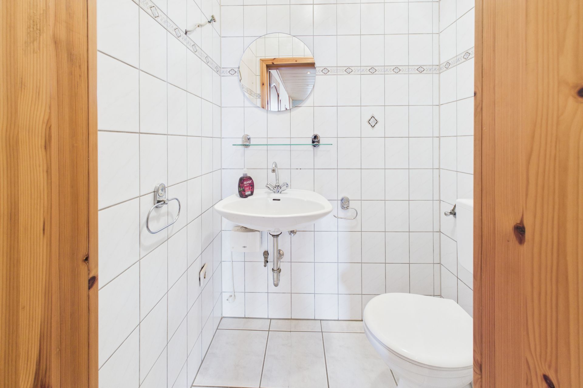 Kleines Badezimmer mit Waschbecken, Spiegel und Toilette, weiße Fliesen an Wänden und Boden.