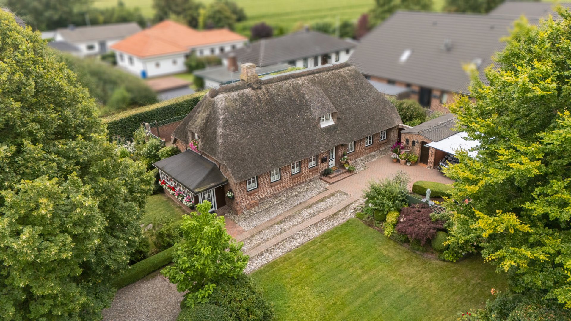 Backsteinhaus mit Reetdach in einem Garten, umgeben von Bäumen und anderen Häusern.