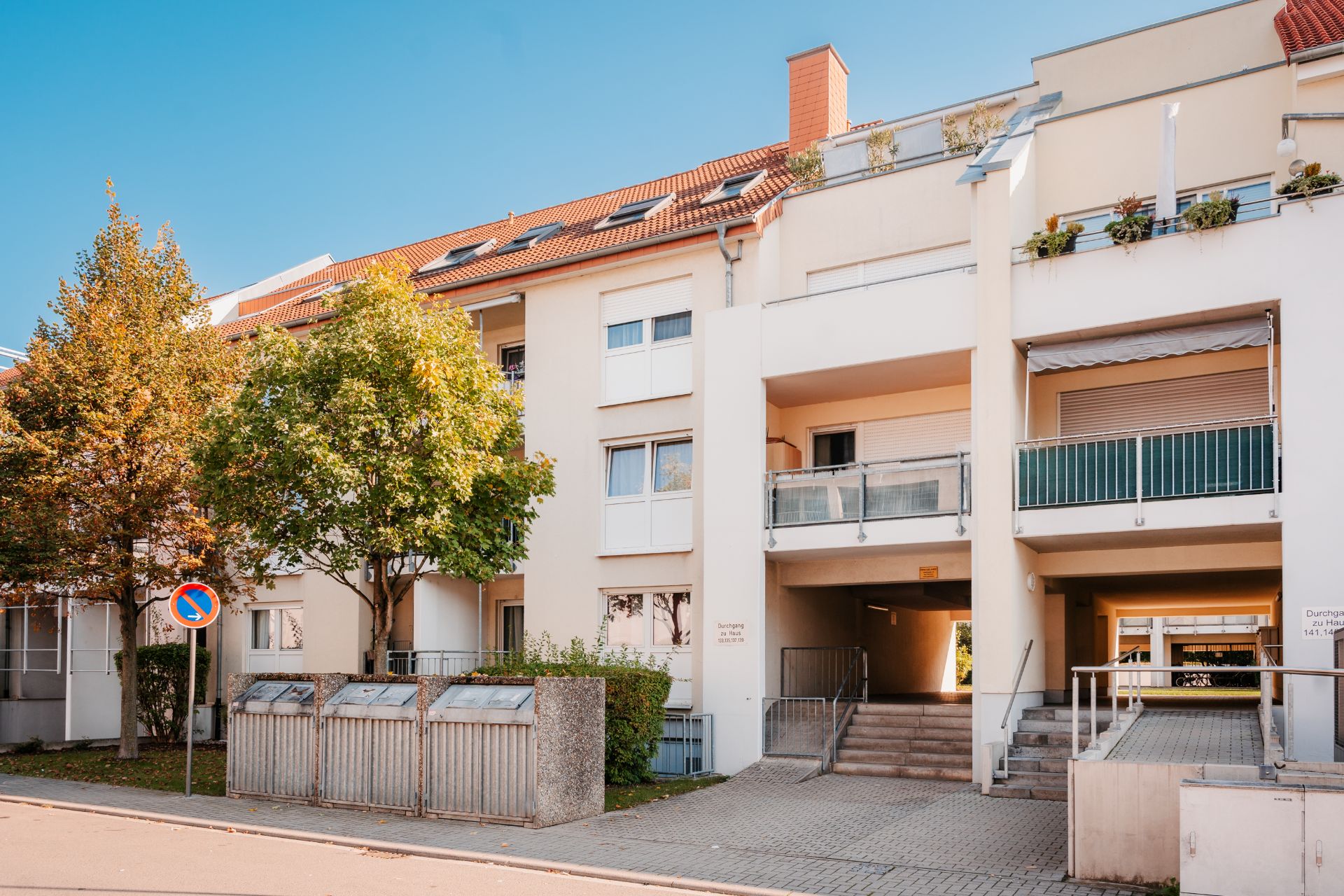 Mehrfamilienhaus mit Balkonen, Treppenaufgang und Mülltonnen am Straßenrand.