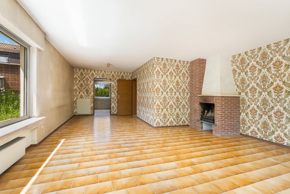 Helles Wohnzimmer mit großem Fenster, gemusterten Tapeten, Kamin und braun gefliestem Boden.