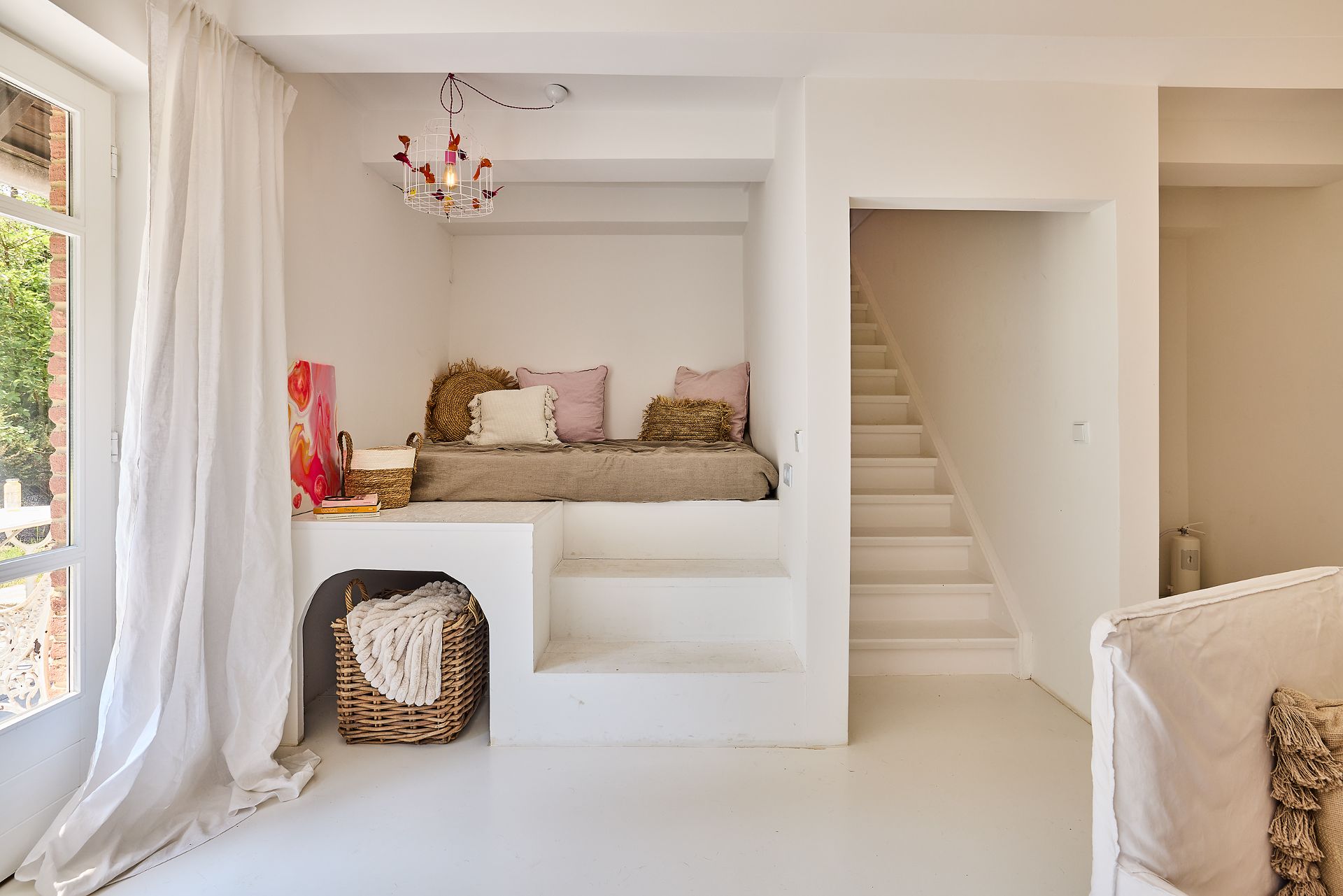 Helles Zimmer mit eingebautem Sofa, weißen Stufen und einem Korb unter der Sitzecke.