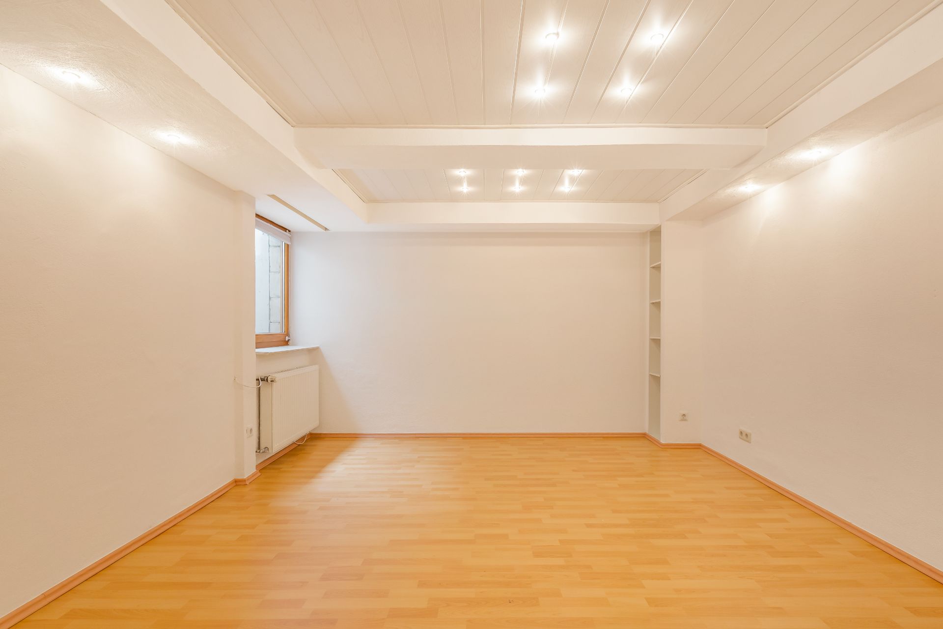 Leerer heller Raum mit Holzboden, weißer Decke, eingebauten Lichtern und einem Fenster.