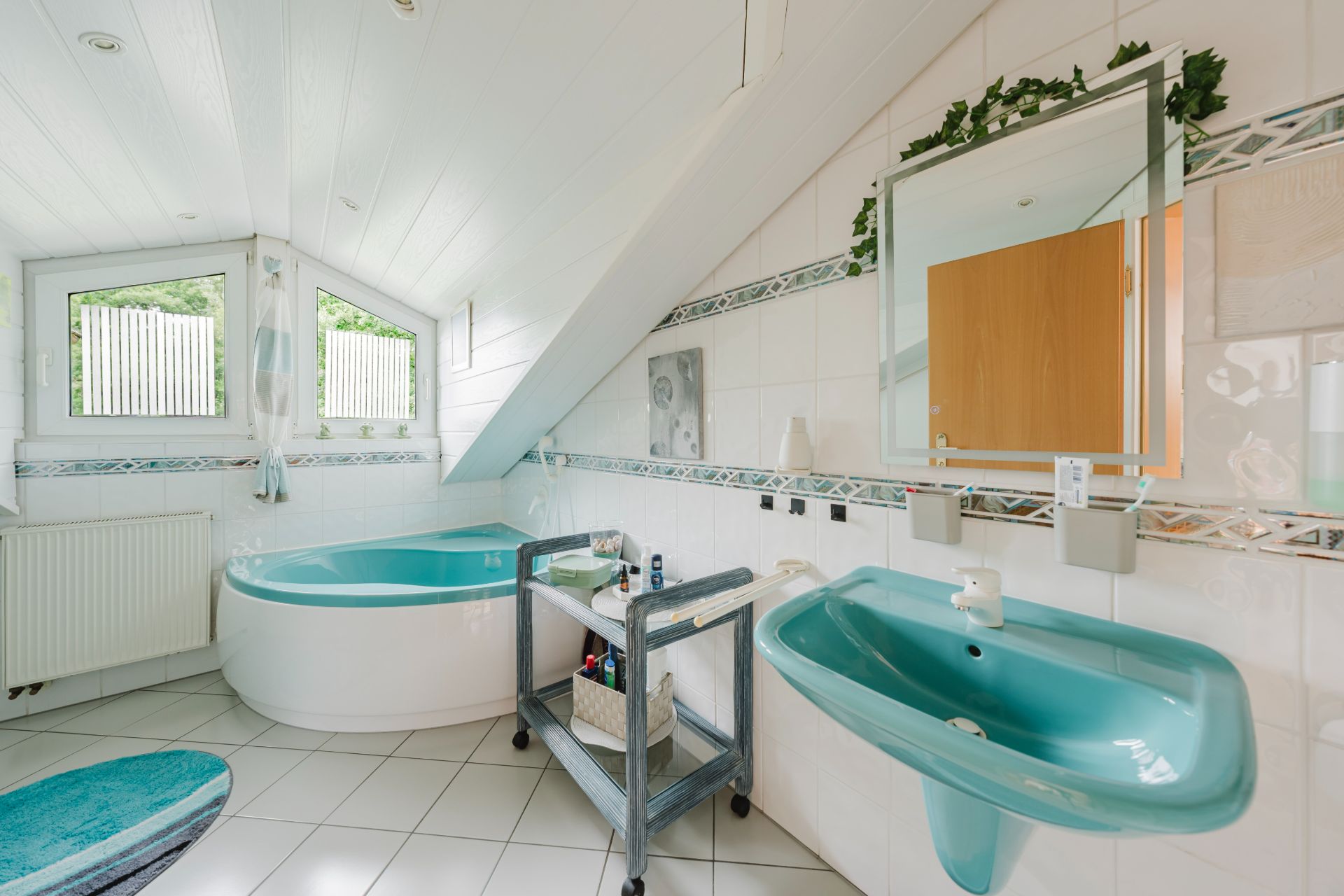 Helles Badezimmer mit Eckbadewanne, türkisfarbenem Waschbecken und zwei Fenstern unter einer Dachschräge.