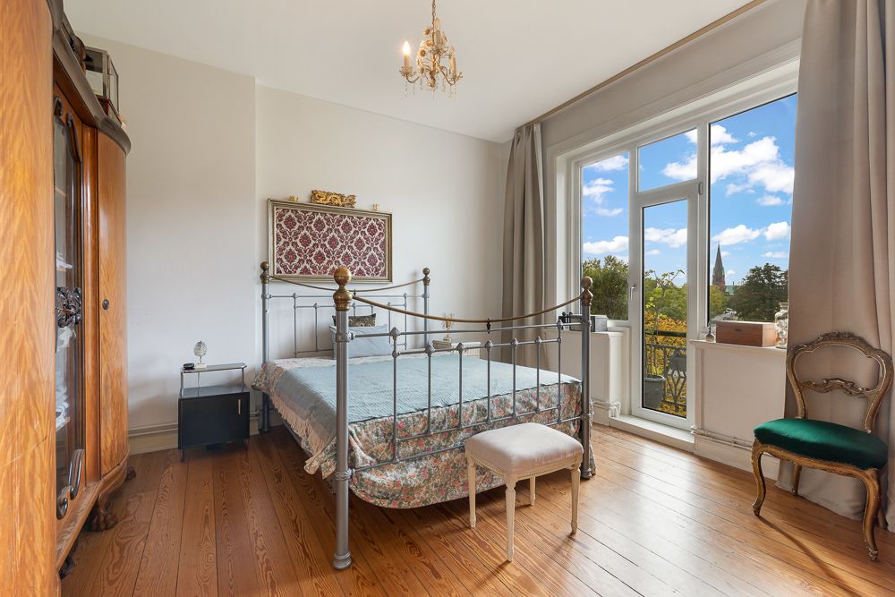 Schlafzimmer mit Metallbett, Holzschrank, Stuhl und großem Fenster mit Blick ins Grüne.
