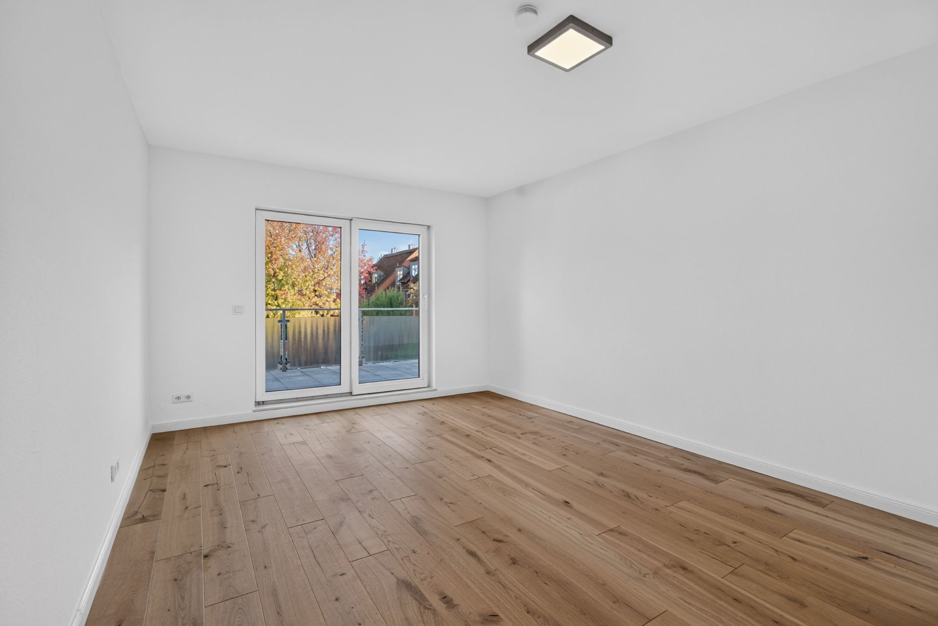 Leerer Raum mit Holzfußboden, weißen Wänden und großer Glastür zum Balkon.