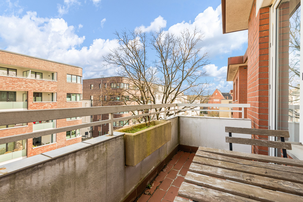 Balkon mit Holztisch und Bank, Blick auf Wohngebäude und Bäume bei Tageslicht.