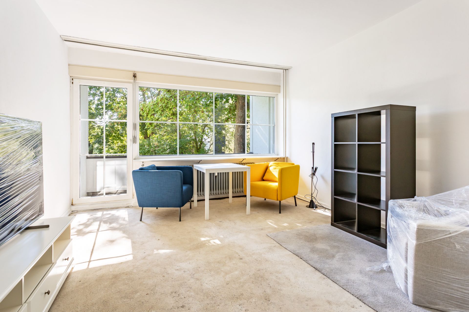 Helles Wohnzimmer mit großen Fenstern, zwei Sesseln, Regal, Tisch und Fernseher.