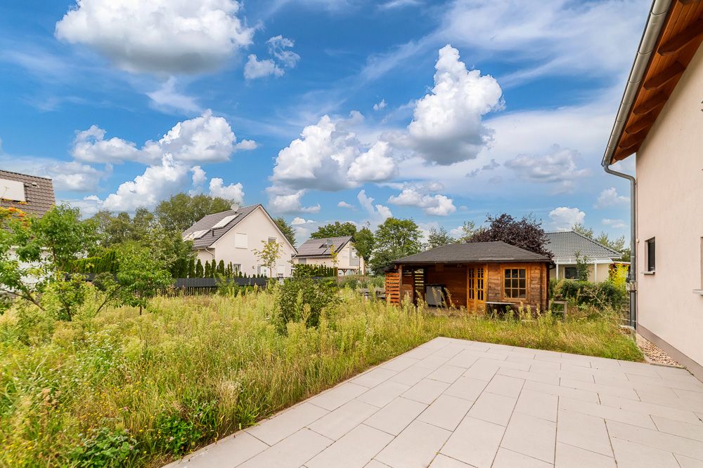 Gepflegter Garten mit Terrasse und Holzhaus, umgeben von weiteren Wohnhäusern bei blauem Himmel.