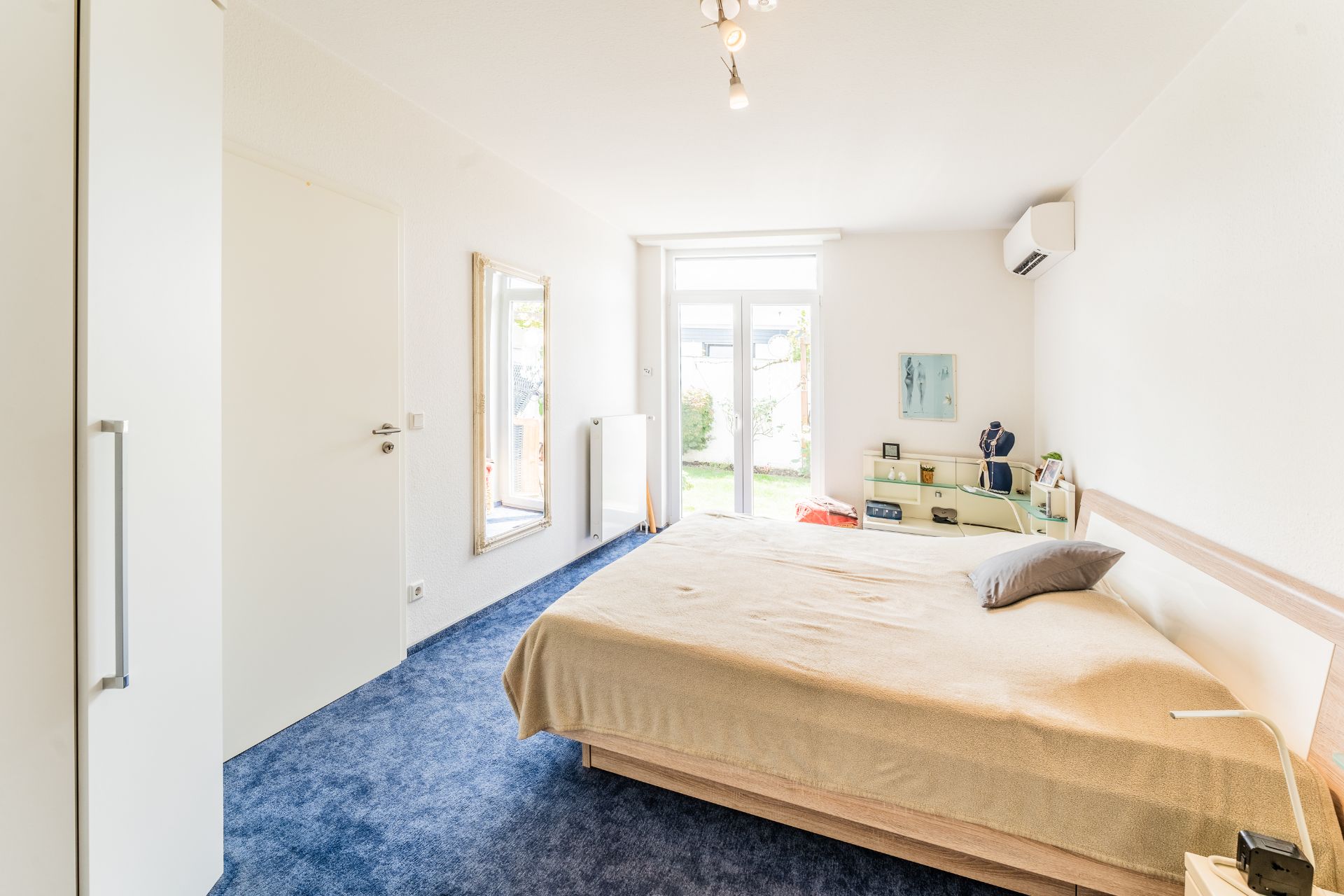 Helles Schlafzimmer mit Doppelbett, blauem Teppich, großem Spiegel und Tür zum Garten.