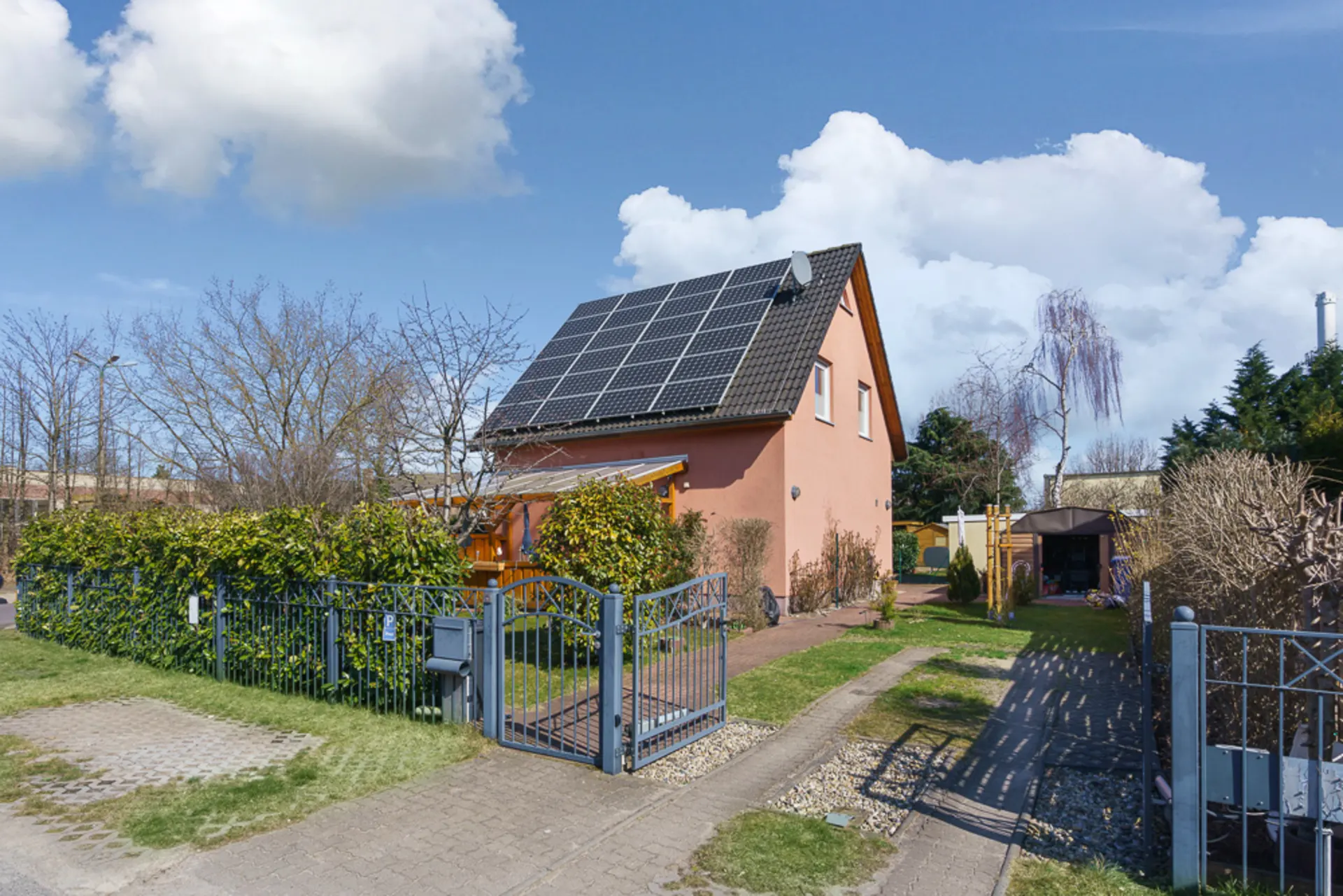 Einfamilienhaus mit Solarpanels auf dem Dach, Garten und Einfahrt, umgeben von Zaun und Büschen.