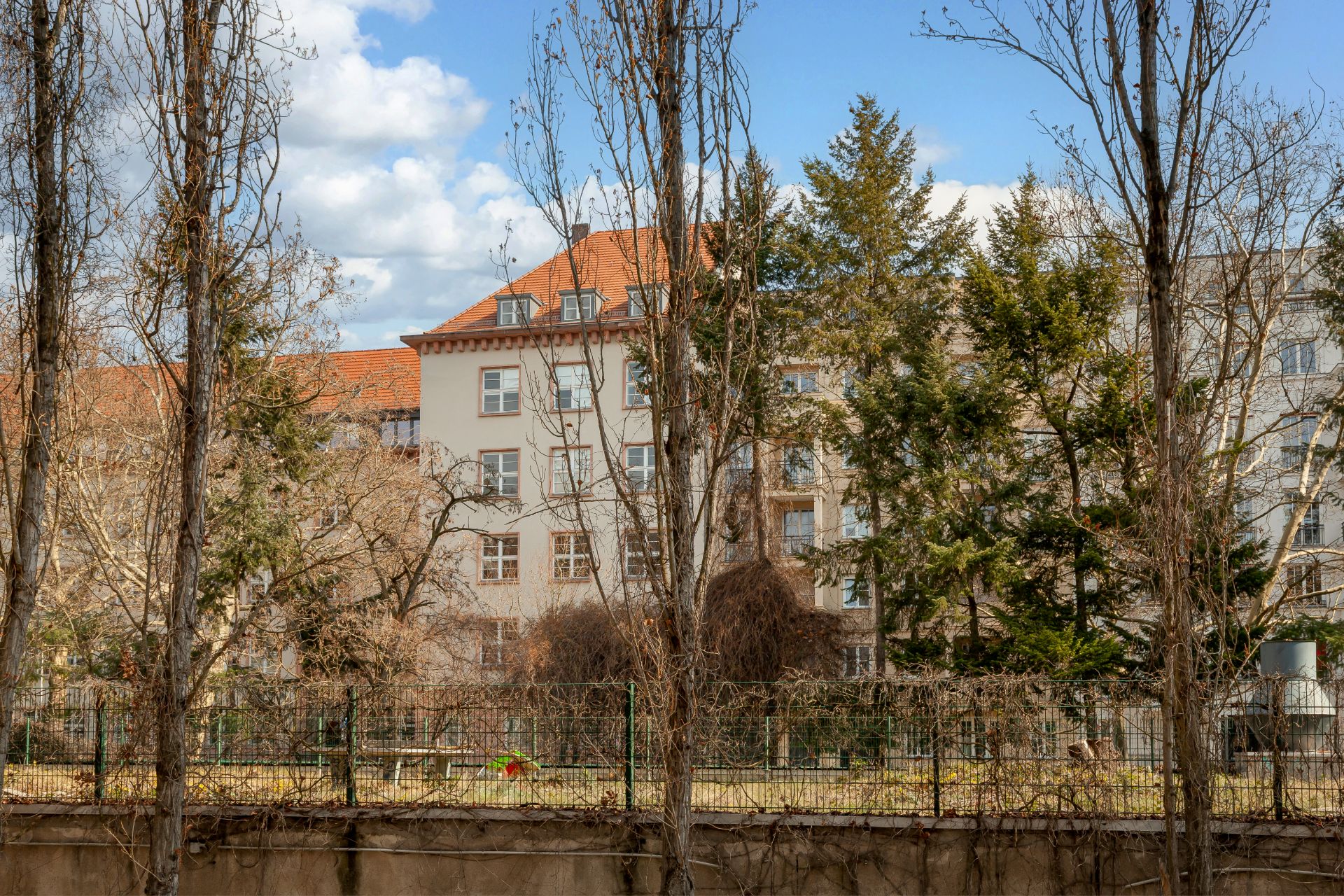 Mehrstöckiges Wohngebäude mit rotem Dach, Bäumen und Garten im Vordergrund.