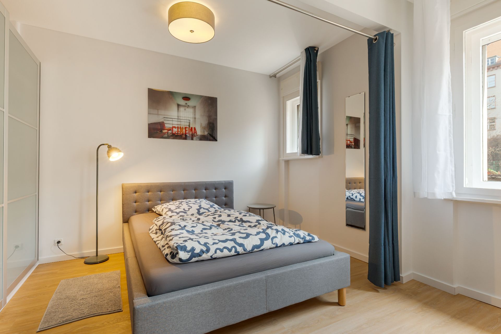 Helles Schlafzimmer mit grauem Bett, Stehlampe, Wandbild und großem Fenster.