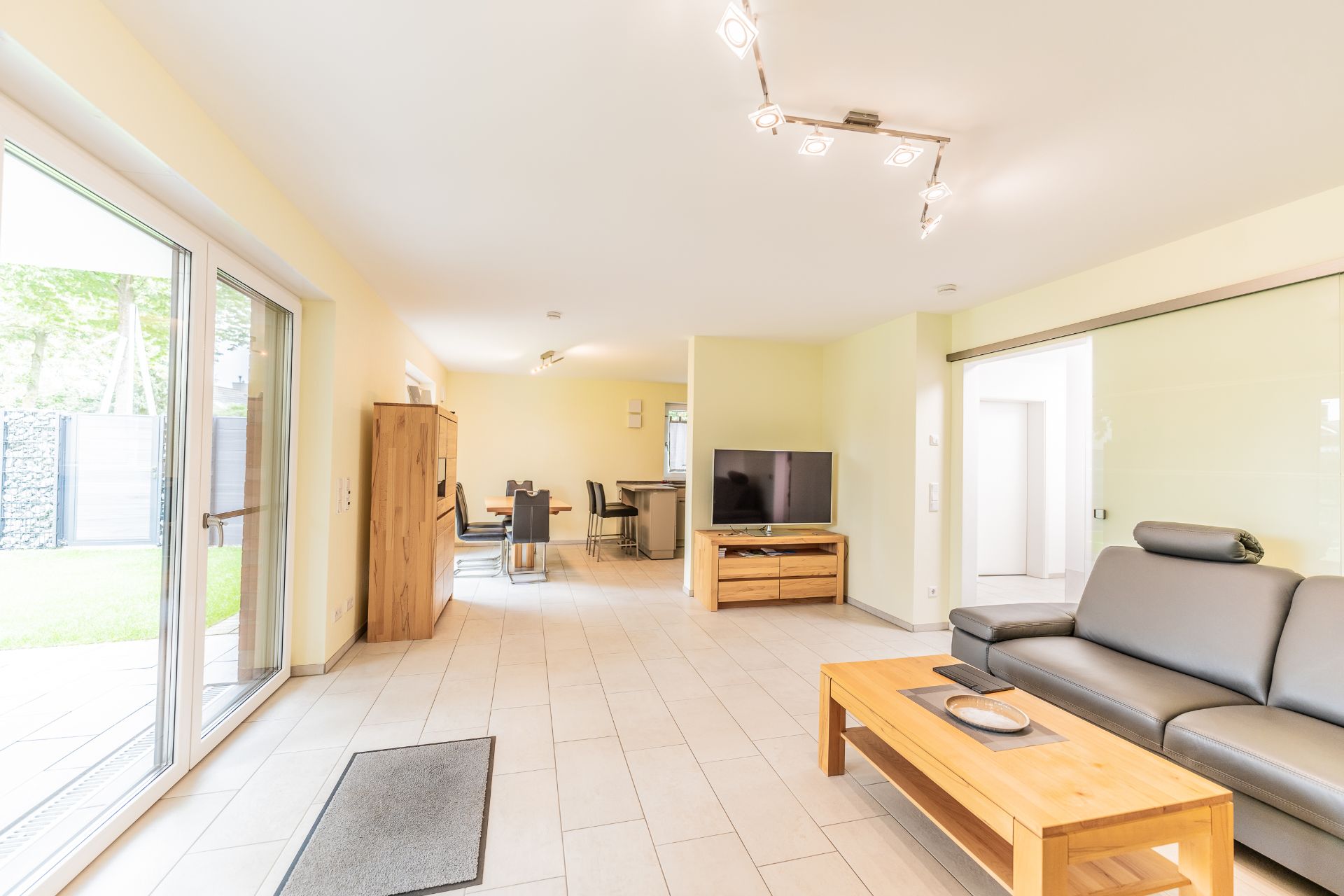 Helles Wohnzimmer mit grauem Sofa, Holztisch, Fernseher und Essbereich. Große Fenster mit Blick auf den Garten.