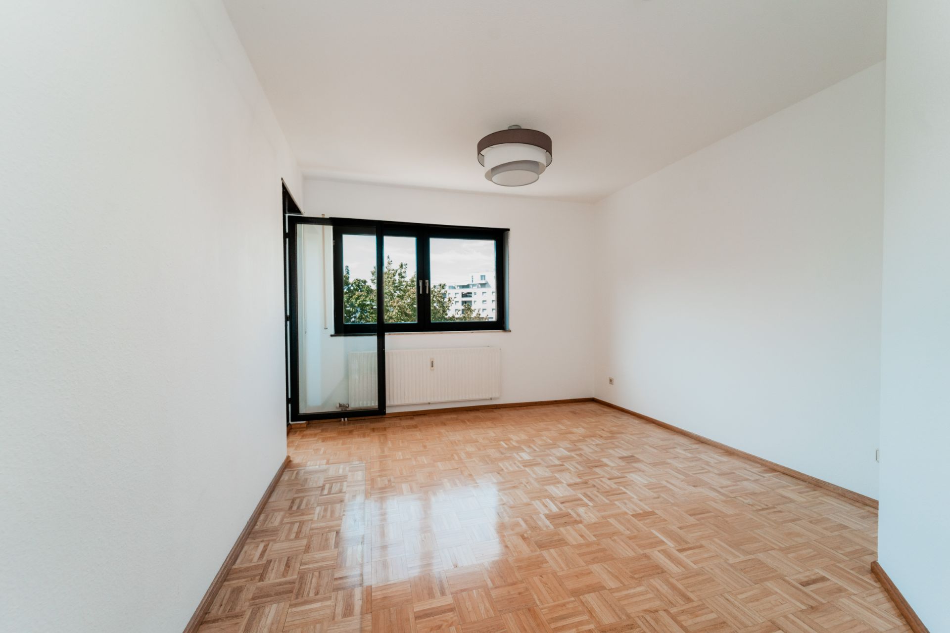 Leeres Zimmer mit Parkettboden, weiß gestrichenen Wänden und großem Fenster mit Blick ins Freie.