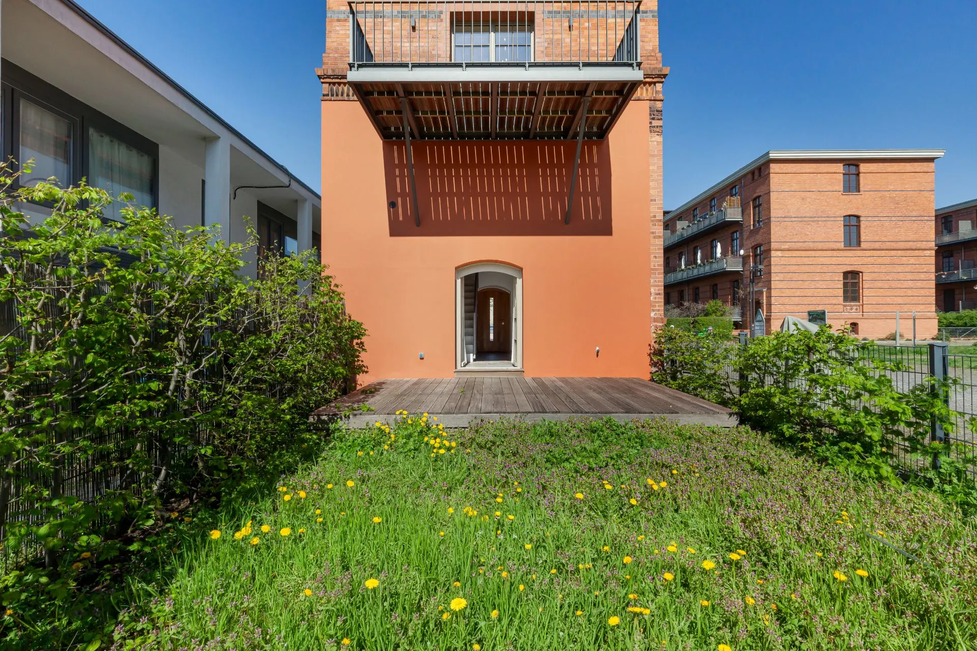 Garten mit Rasen und gelben Blumen vor einem roten Wohnhaus mit Terrasse und Balkon.