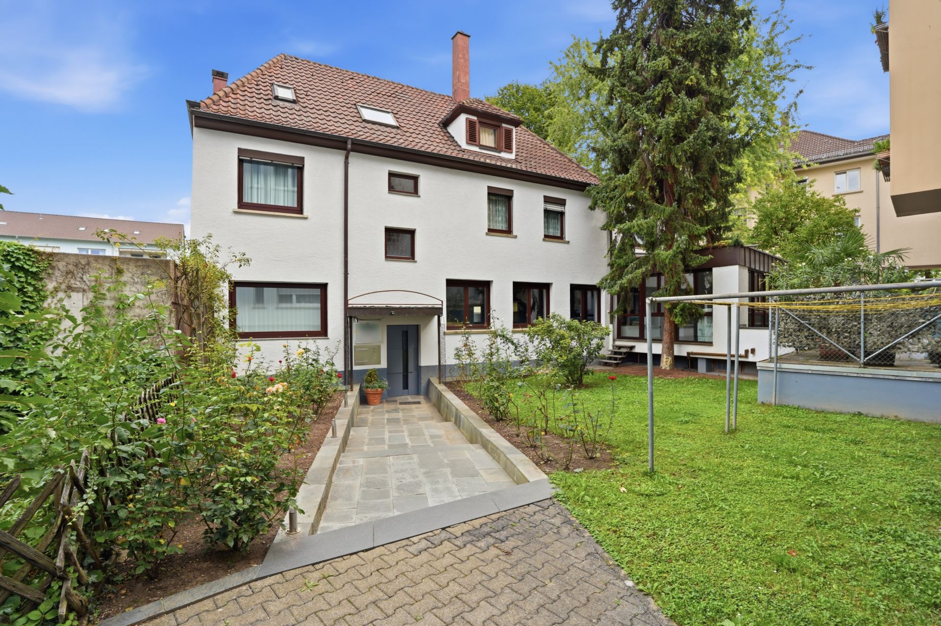 Zweistöckiges Wohnhaus mit Garten, Gehweg und Bäumen im Vordergrund.