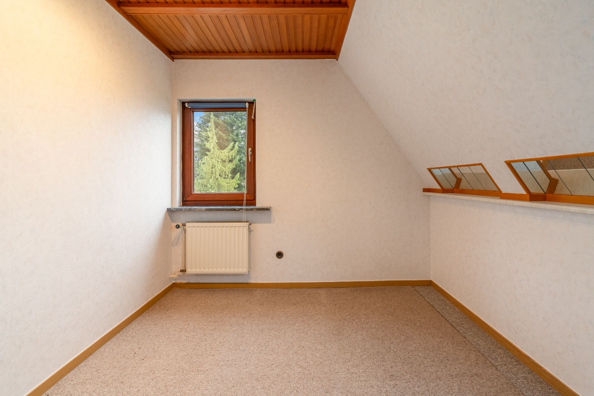 Leerer Raum mit Fenster, Heizkörper und schräger Decke mit Holzverkleidung.