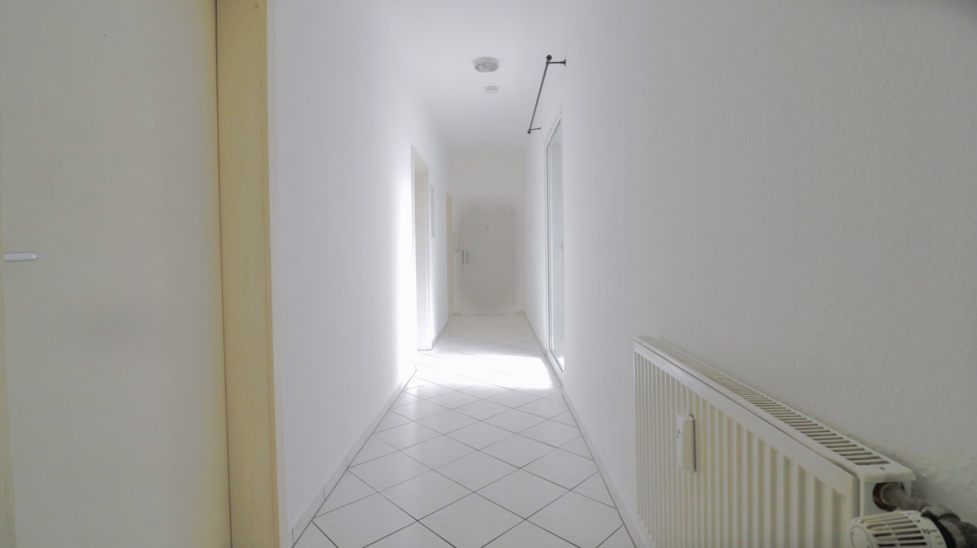 Heller Flur mit weißen Wänden, Fliesenboden und einem Heizkörper an der rechten Wand.