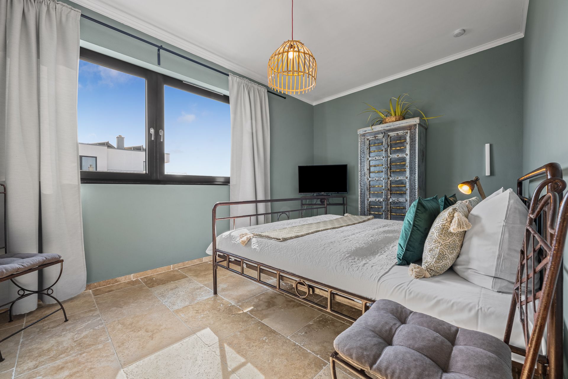 Helles Schlafzimmer mit Metallbett, großem Fenster, Kommode und Fliesenboden.