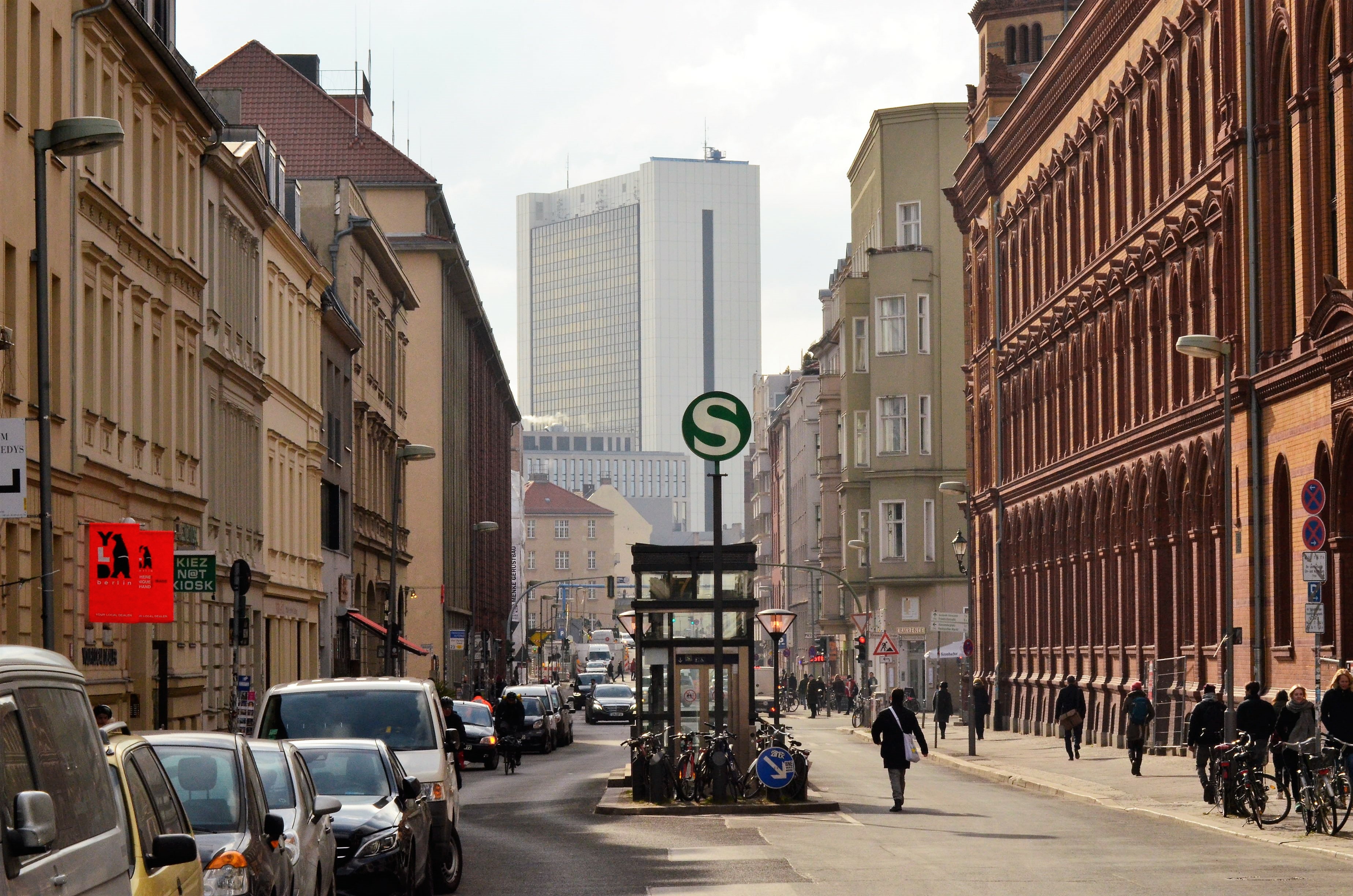 Stadtstraße mit parkenden Autos, Fahrrädern, Menschen und S-Bahn-Zeichen.