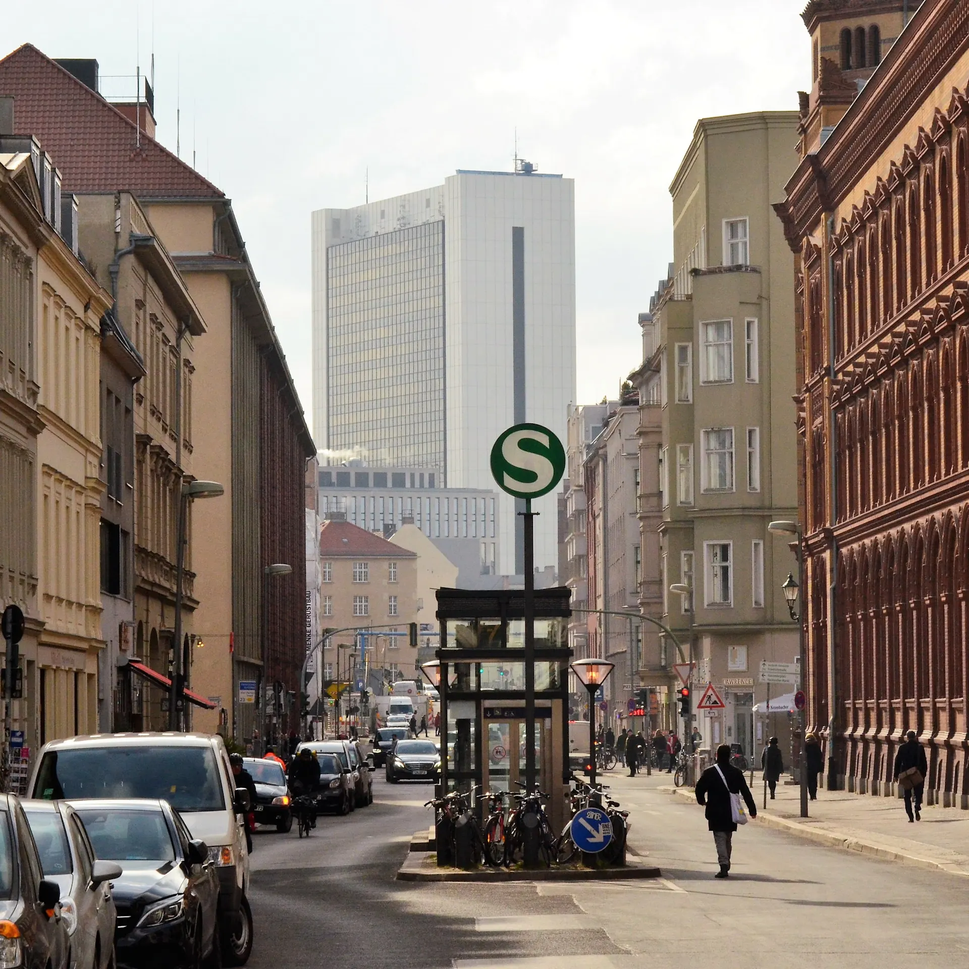 Stadtstraße mit parkenden Autos, Fahrrädern, Menschen und S-Bahn-Zeichen.