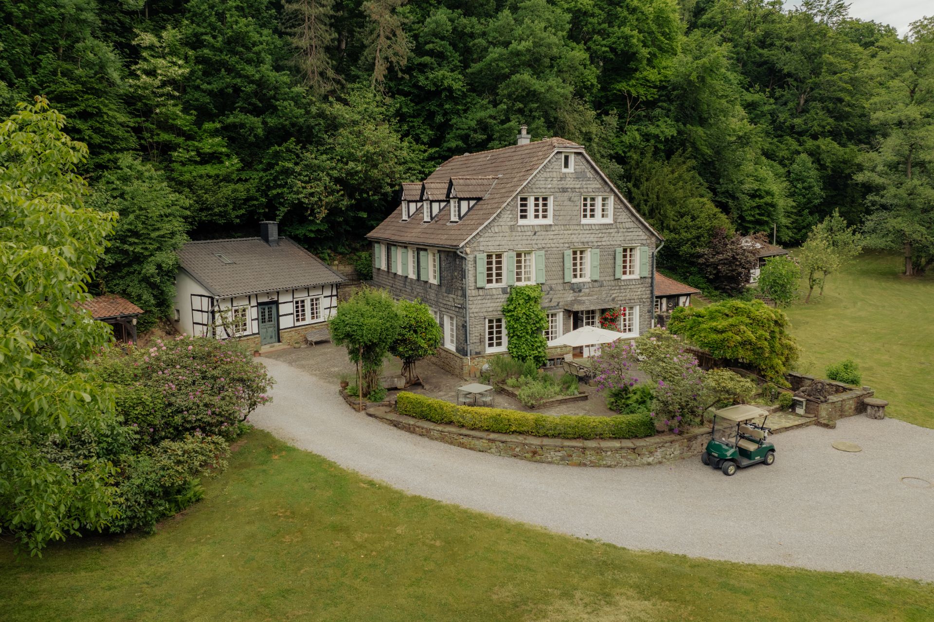 Großes Wohnhaus mit Garten, Auffahrt und Nebengebäude, umgeben von Bäumen und Rasen.