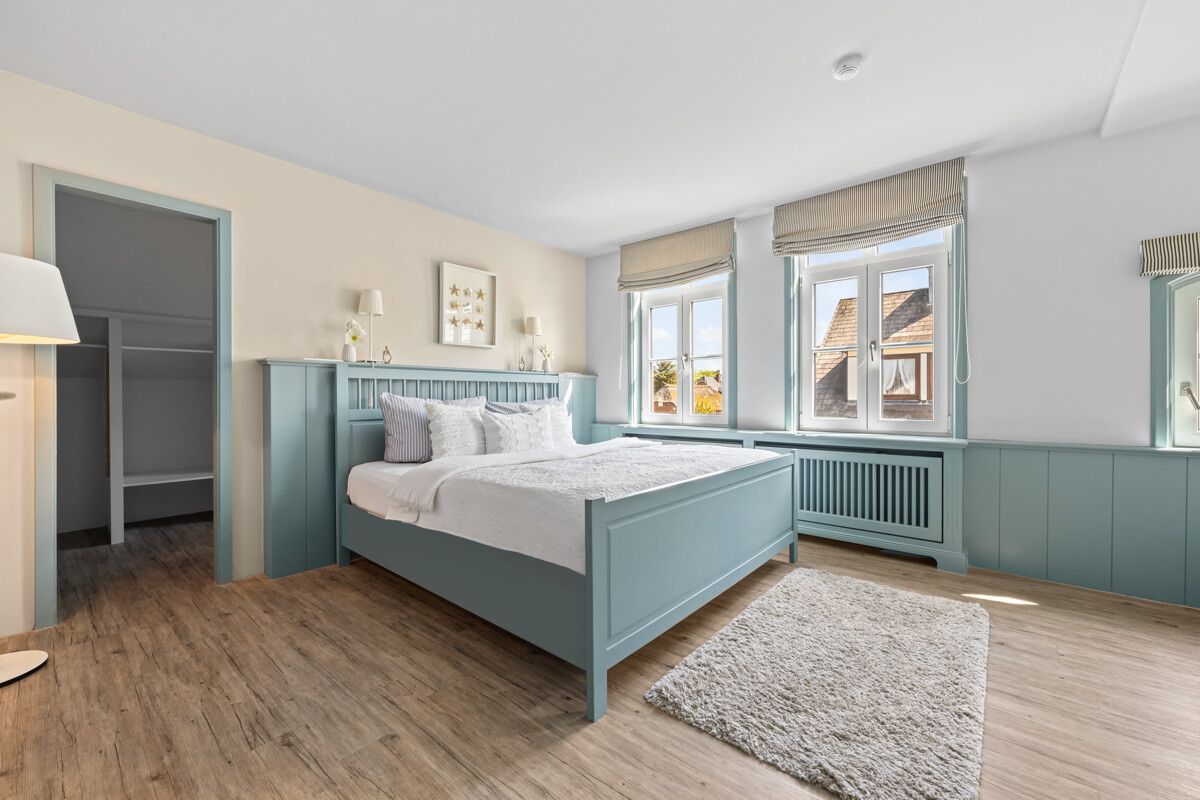 Helles Schlafzimmer mit blauem Bett, Holzfußboden, Teppich und drei Fenstern.
