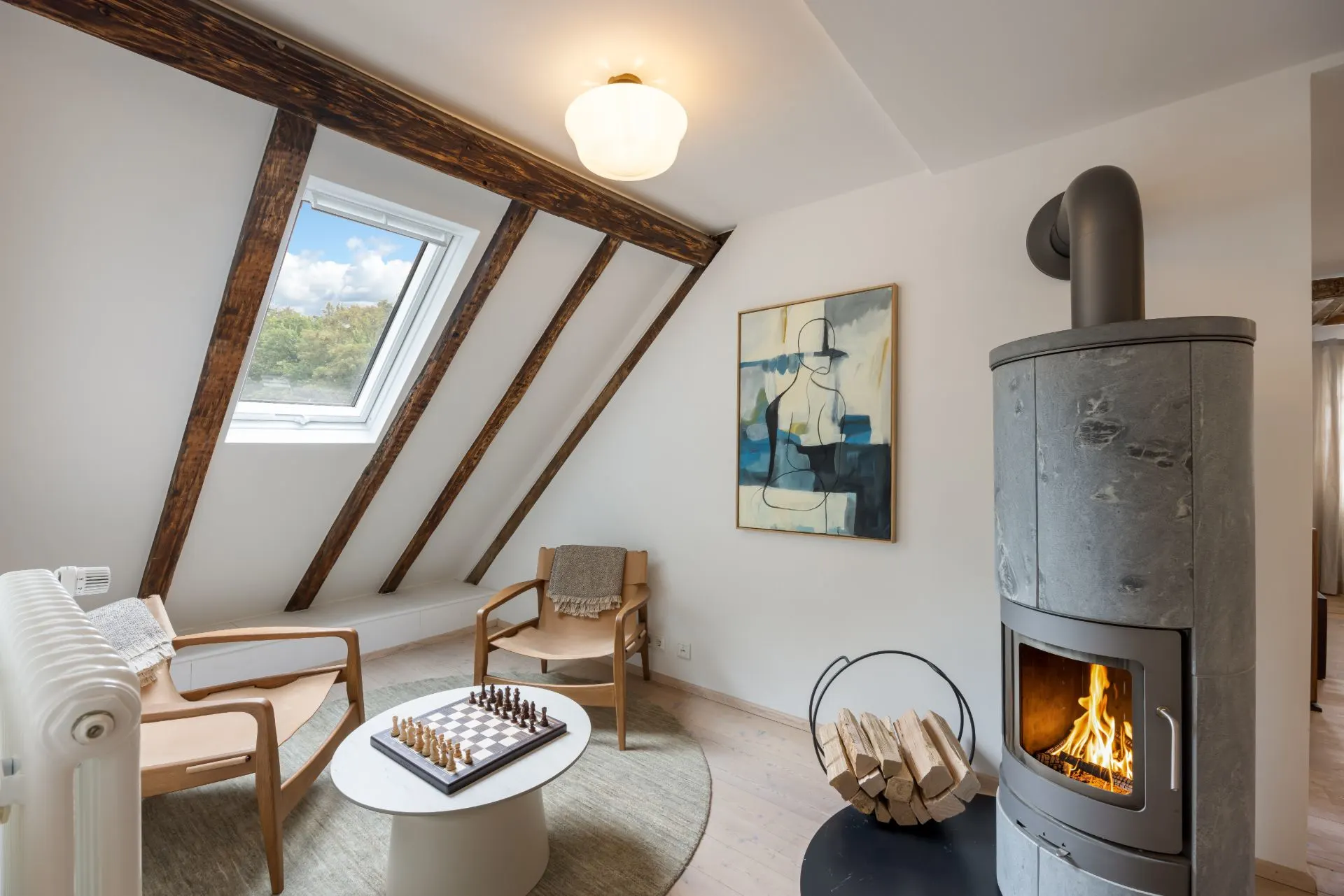 Gemütliches Wohnzimmer mit Kamin, zwei Sesseln, Schachbrett und großem Fenster im Dachgeschoss.