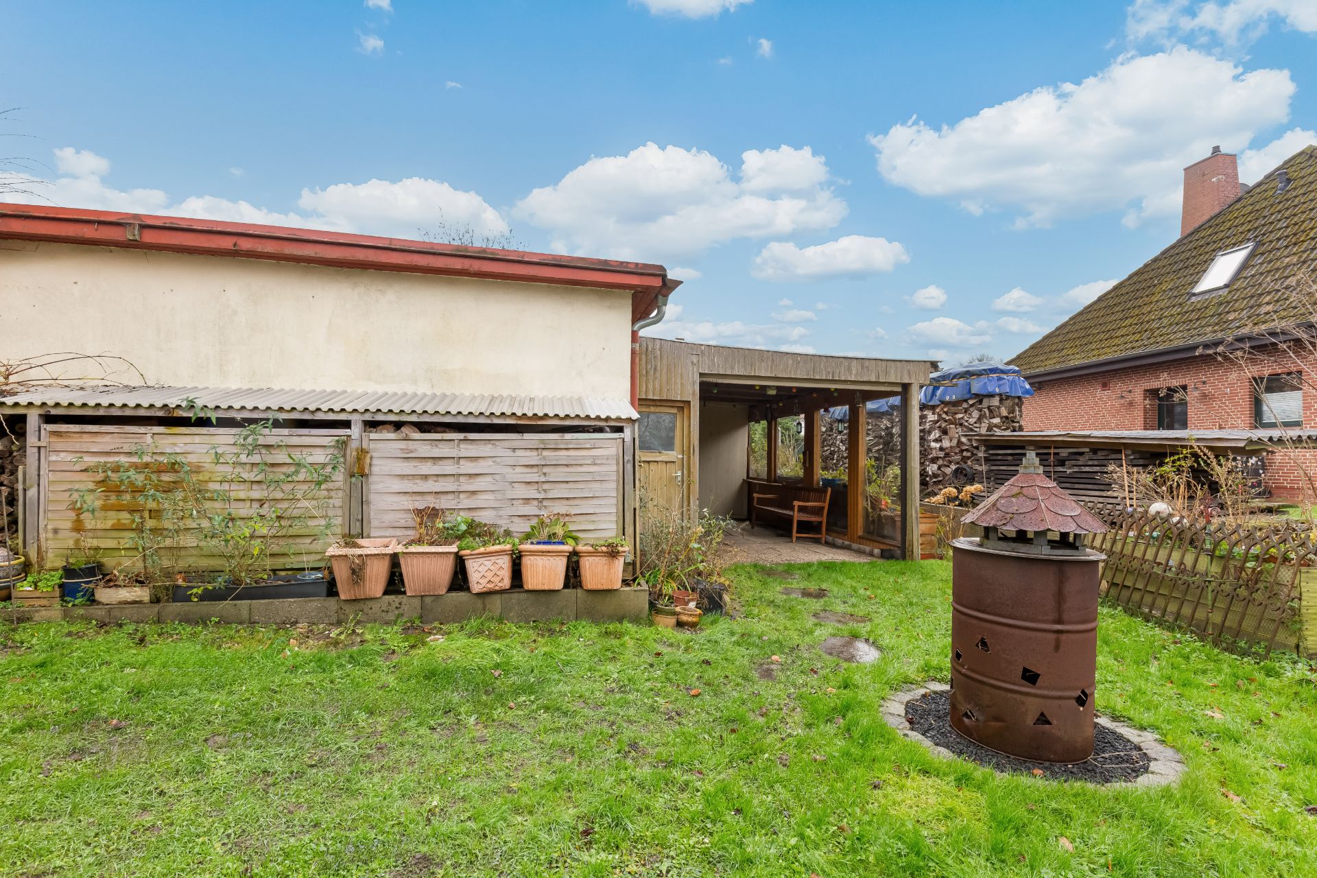 Garten mit Rasenfläche, Blumentöpfen, überdachtem Sitzbereich und Feuerstelle.