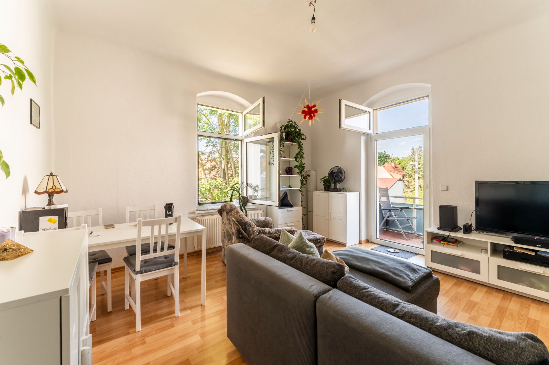 Helles Wohnzimmer mit grauem Sofa, Esstisch, Fernseher und Zugang zu Balkon durch offene Fenster.