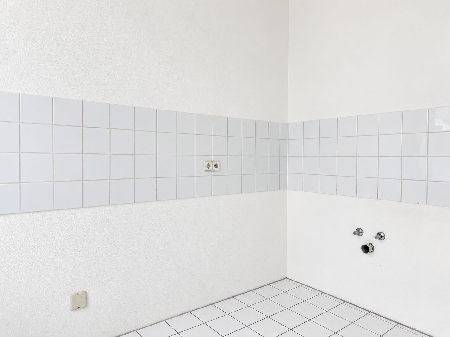 Leere, weiß geflieste Ecke mit Steckdosen und Wasseranschlüssen an der Wand.