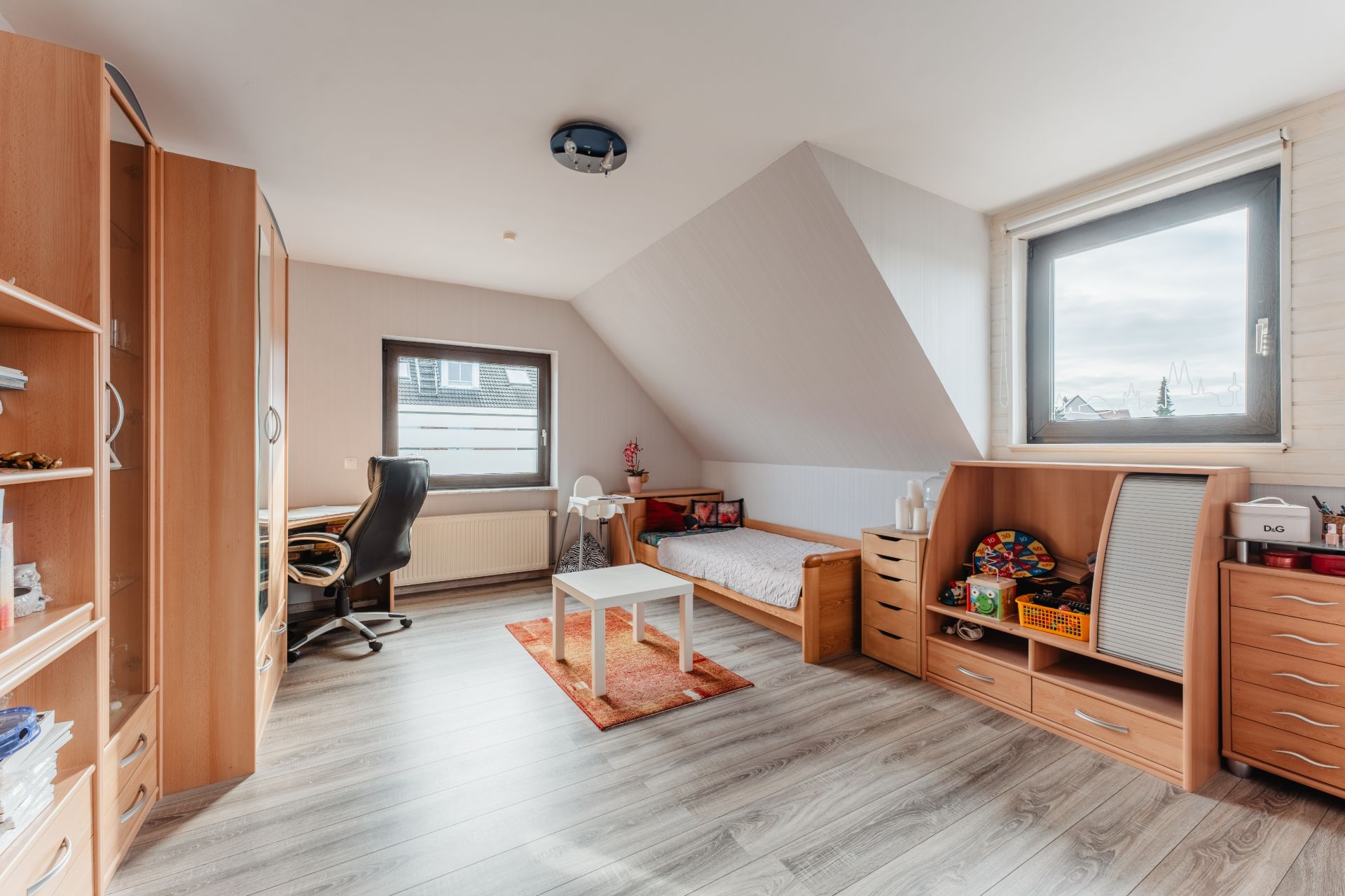 Helles Zimmer mit Holzmöbeln, Schreibtisch, Einzelbett und zwei Fenstern auf Dachgeschoss-Ebene.