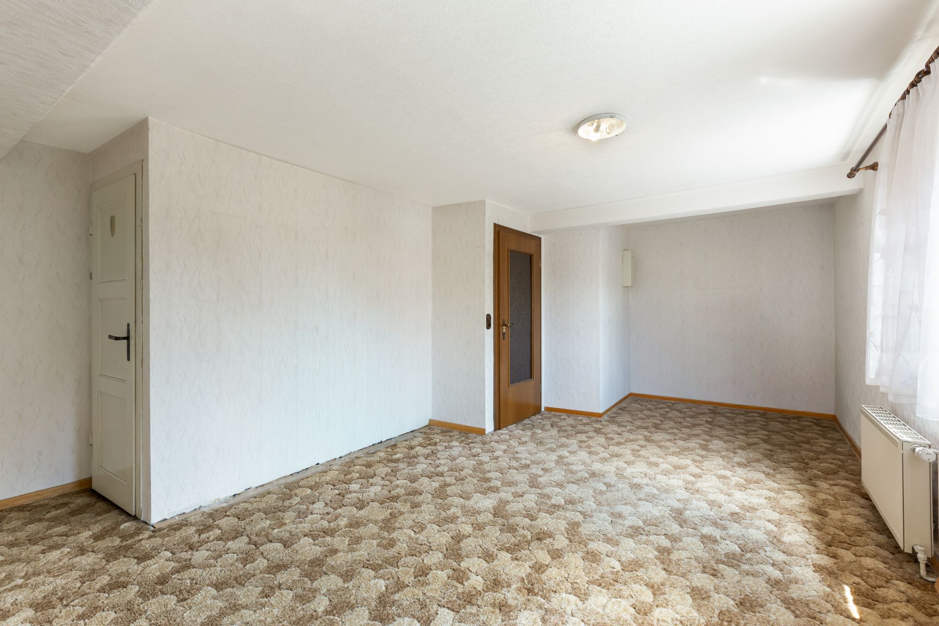 Leeres Zimmer mit Teppichboden, weißer Wand, zwei Türen und großem Fenster mit Vorhängen.