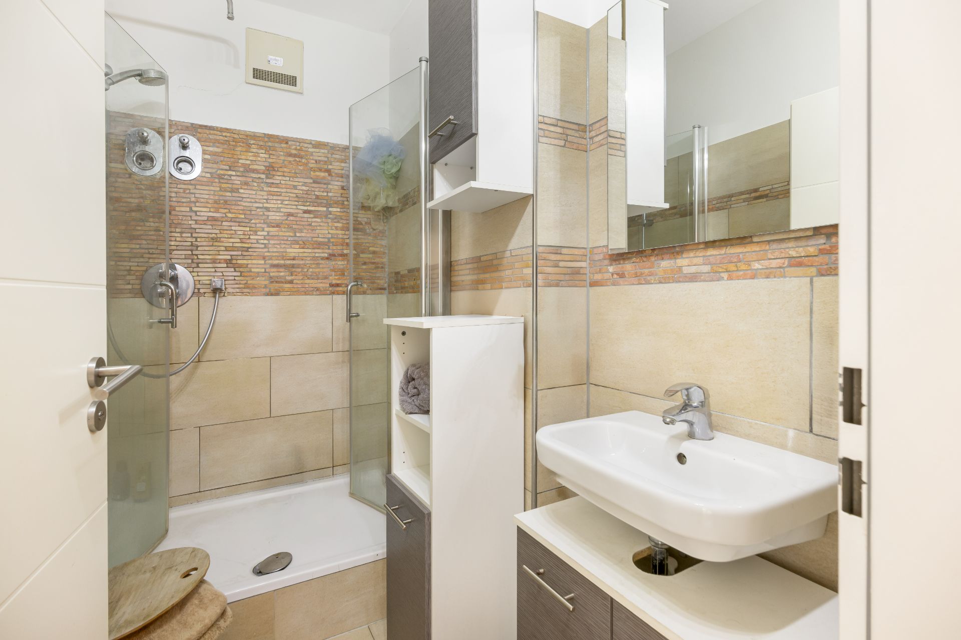 Kleines Badezimmer mit Dusche, Waschbecken, Spiegel und Schrank auf beigen Fliesen.