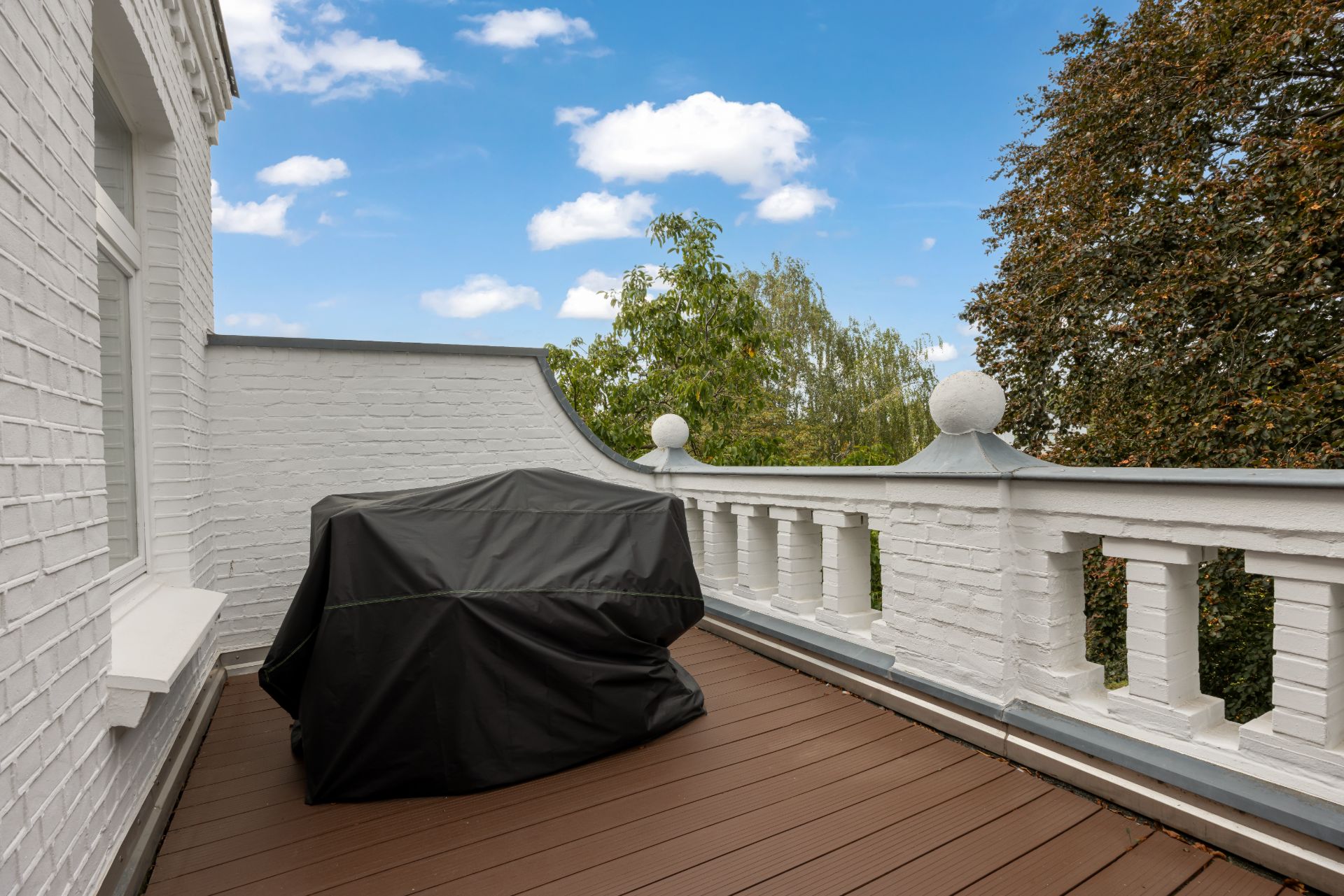 Balkon mit weißer Brüstung, Holzboden und abgedecktem Möbelstück, Bäume im Hintergrund.
