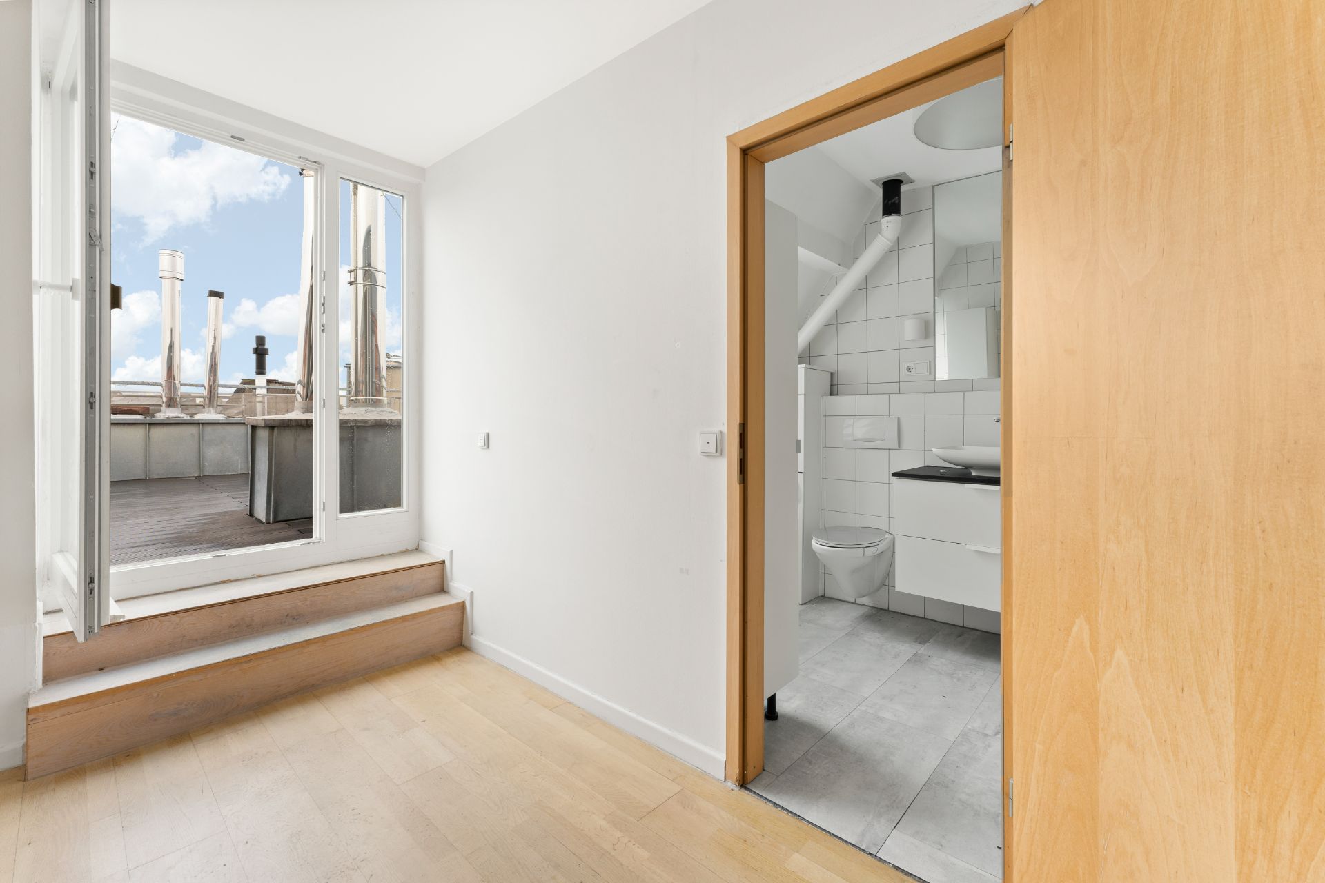 Heller Raum mit Holzboden, Zugang zu Balkon und Blick ins Badezimmer mit weißen Fliesen.
