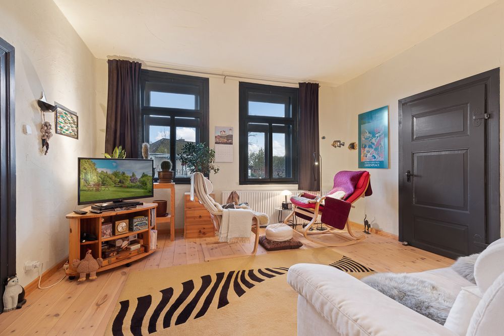 Wohnzimmer mit zwei Sesseln, Fernseher auf Holzregal, Teppich und drei Fenstern mit dunklen Vorhängen.