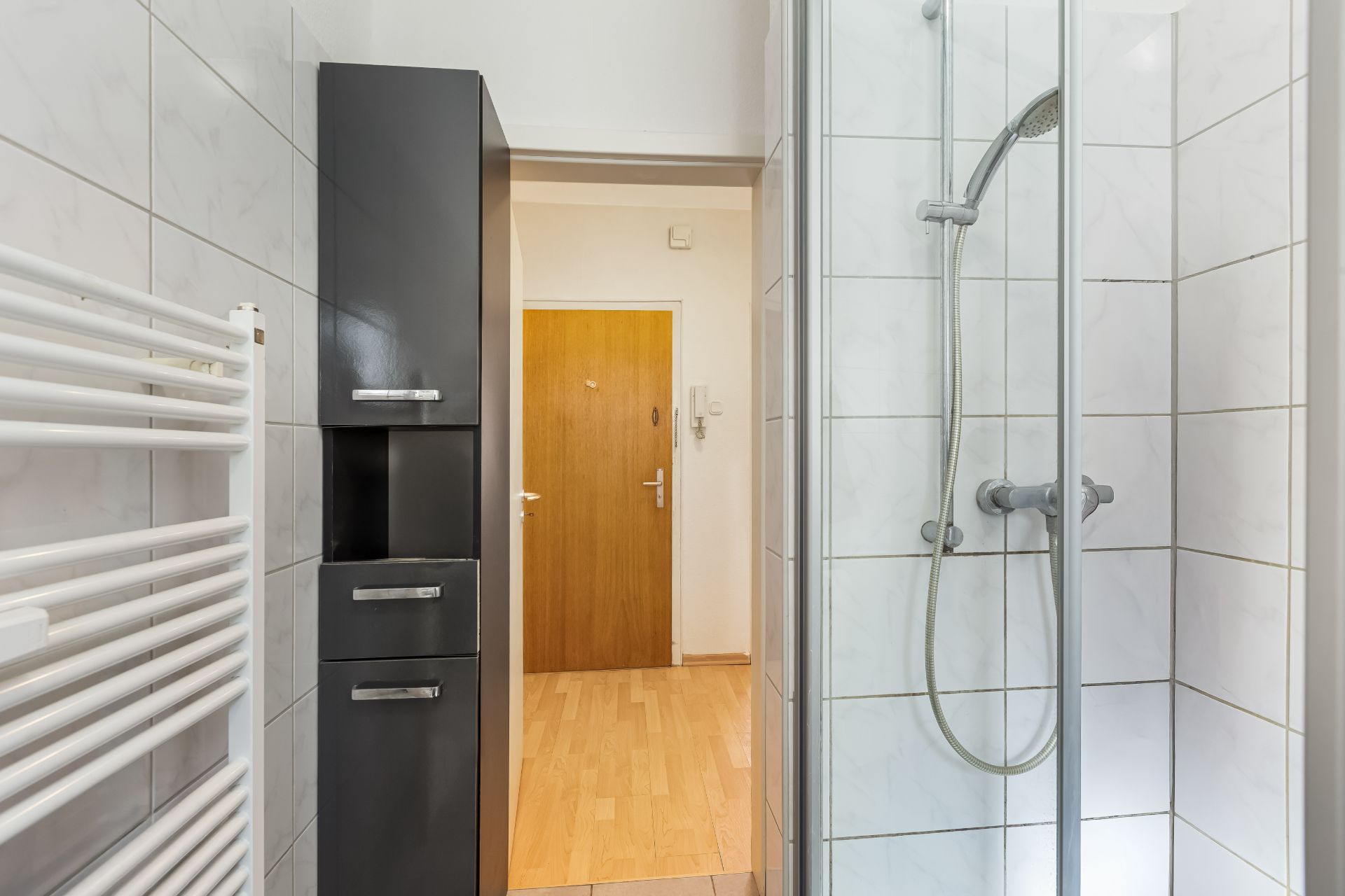 Modernes Badezimmer mit Dusche, Heizkörper, Schrank und Blick auf einen Flur mit Holzboden.