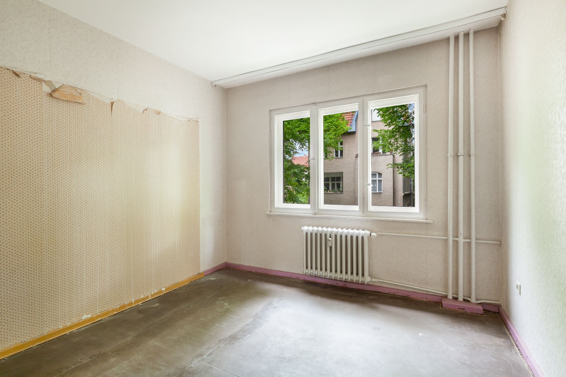 Leeres Zimmer mit großem Fenster, Heizung und teilweise abgerissener Tapete.