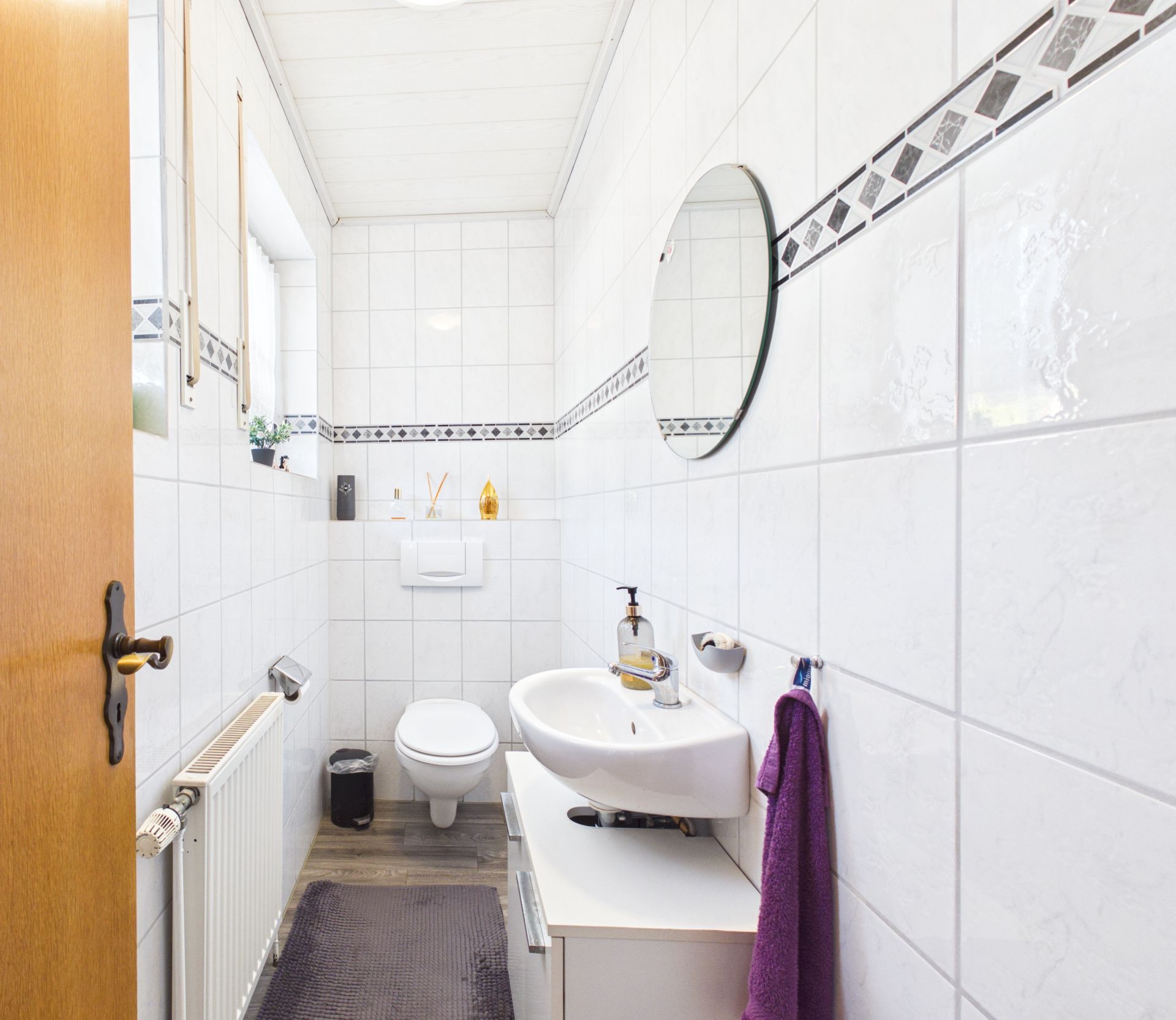 Helles, schmal geschnittenes Badezimmer mit Wandtoilette, Waschbecken und rundem Spiegel.