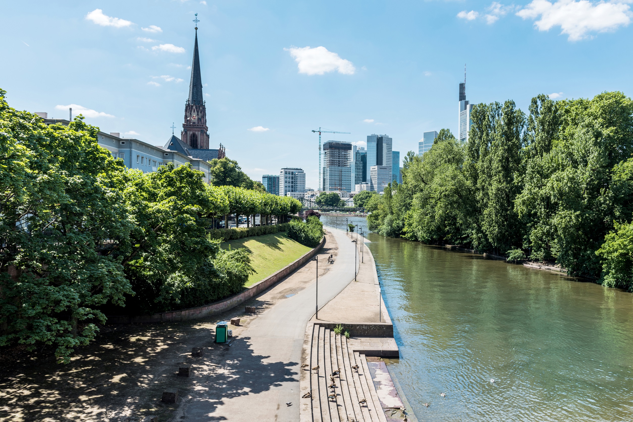Promenade mit Bäumen am Fluss, Kirche und moderne Hochhäuser im Hintergrund.