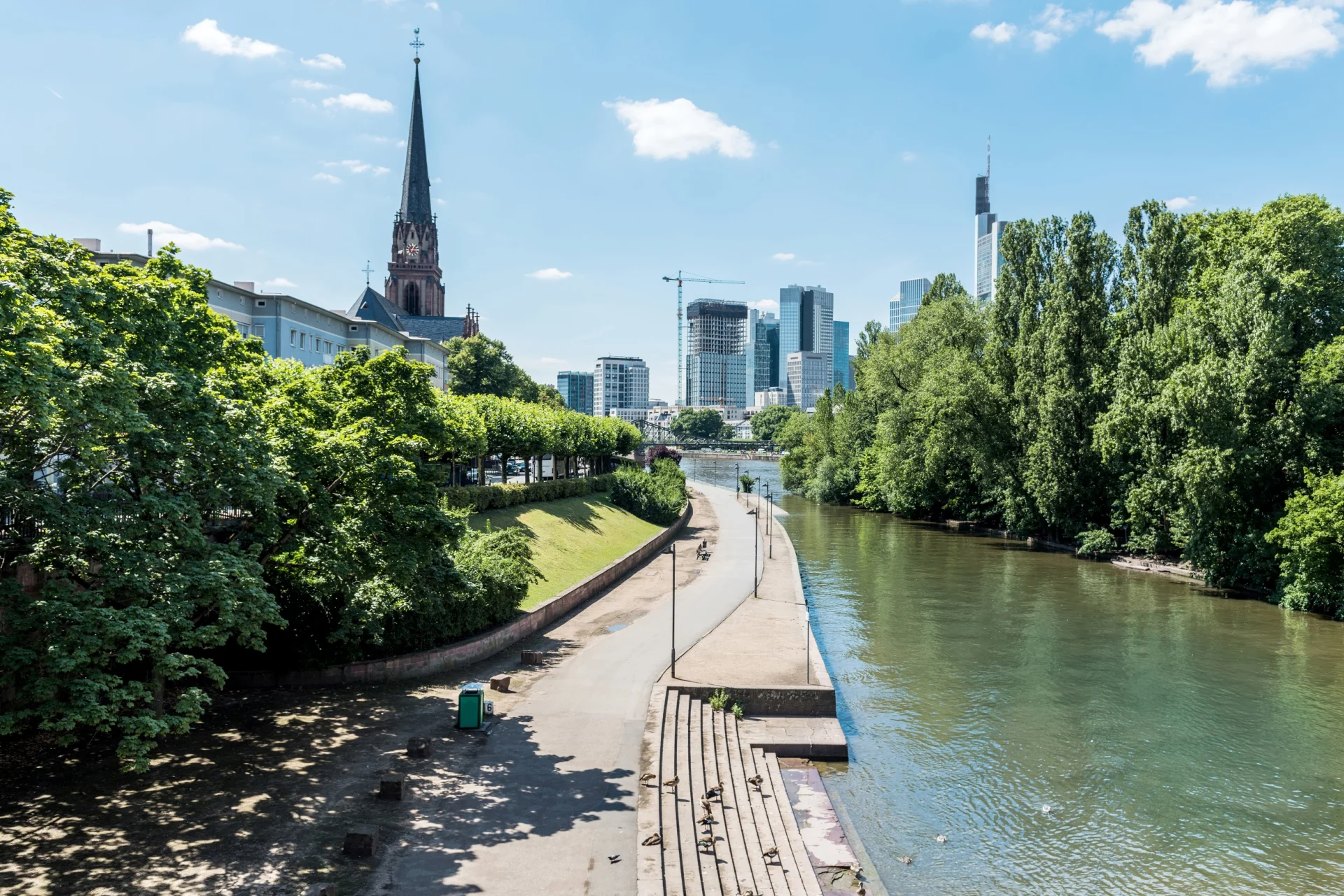 Promenade mit Bäumen am Fluss, Kirche und moderne Hochhäuser im Hintergrund.