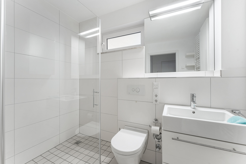 Modernes, weißes Badezimmer mit Dusche, WC, Waschbecken und großem Spiegel.