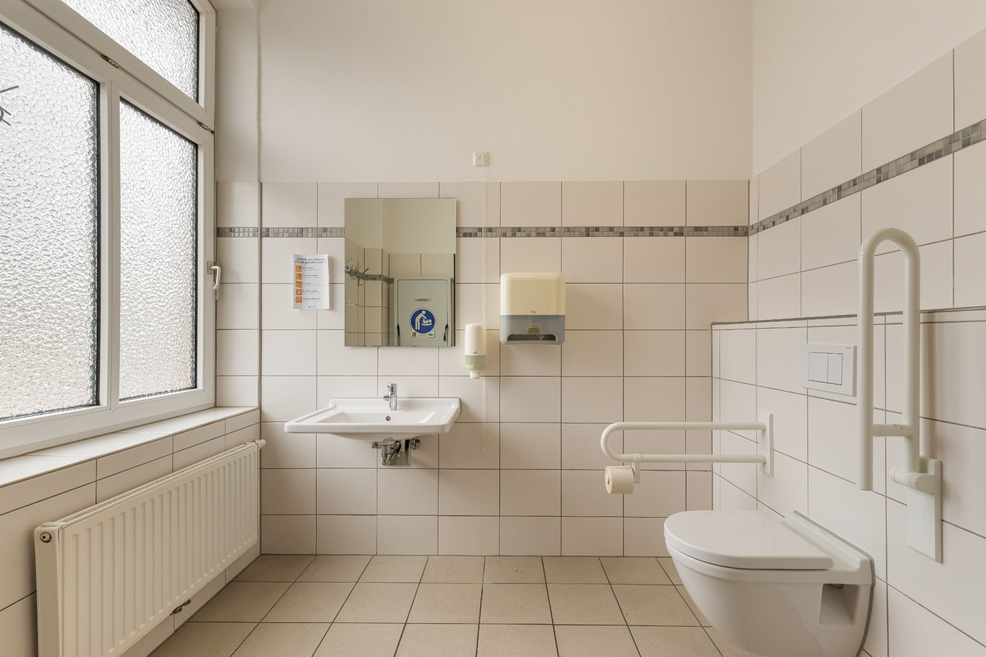 Barrierefreies Badezimmer mit Waschbecken, Spiegel, WC und Haltegriffen.