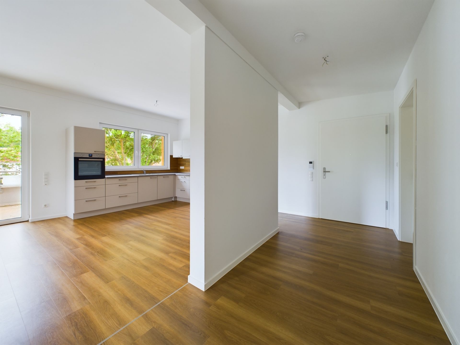 Helles Zimmer mit Parkettboden, offener Küche und großen Fenstern, Blick auf Haustür.