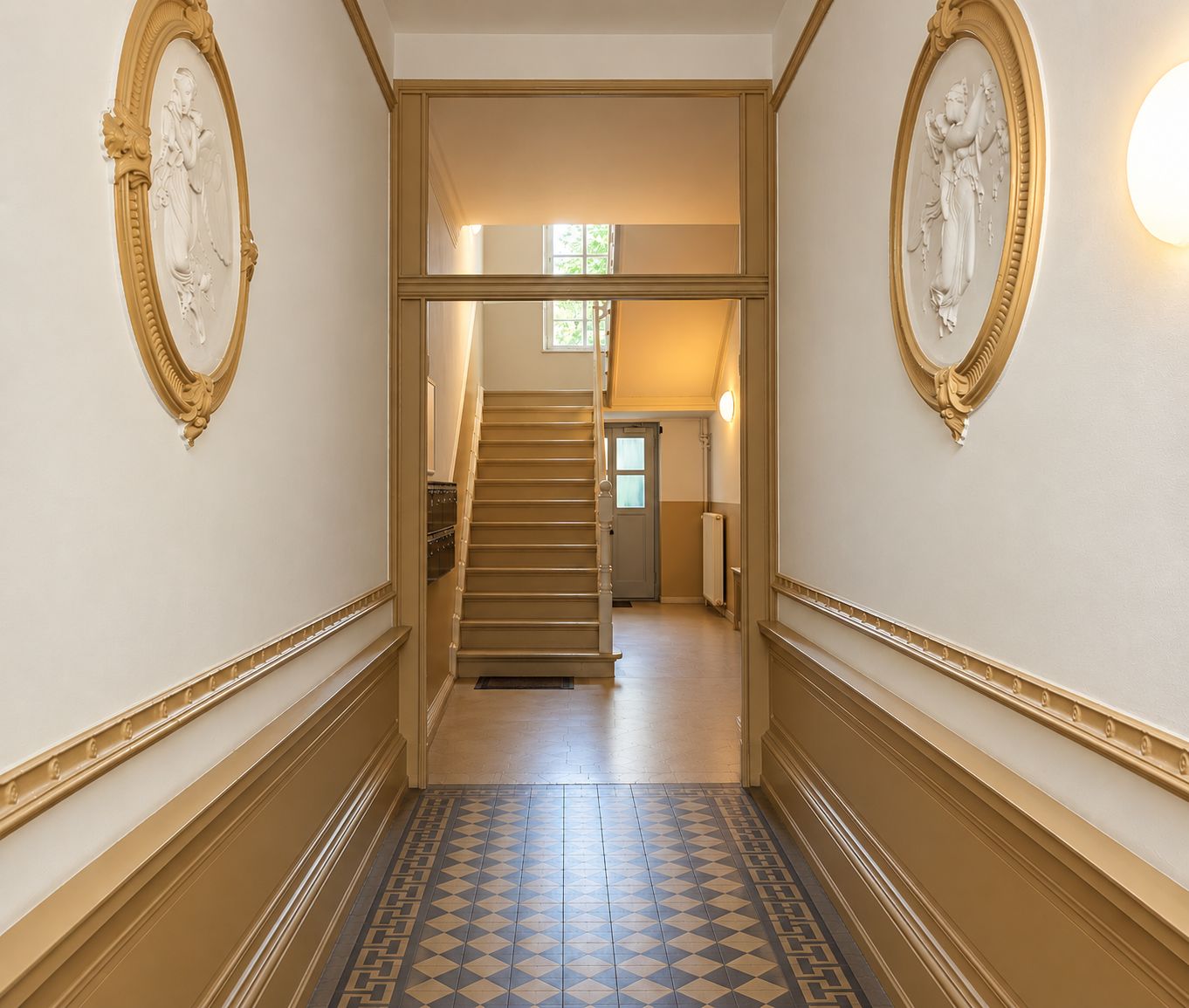 Flur mit gemustertem Boden, Stuckverzierungen an den Wänden und Treppe im Hintergrund.