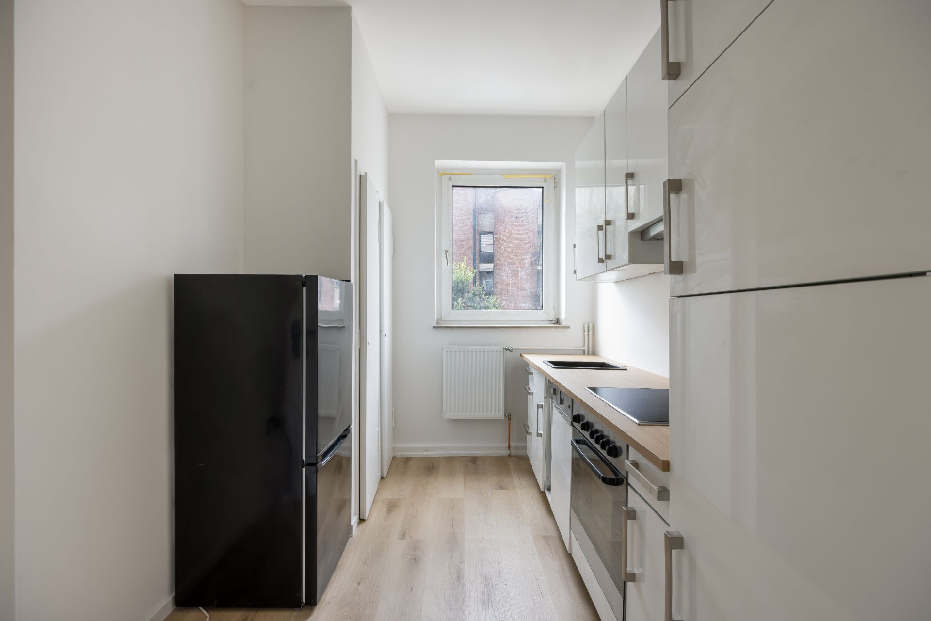 Schmale Küche mit weißem Schrank, schwarzem Kühlschrank und Fenster am Ende des Raums.