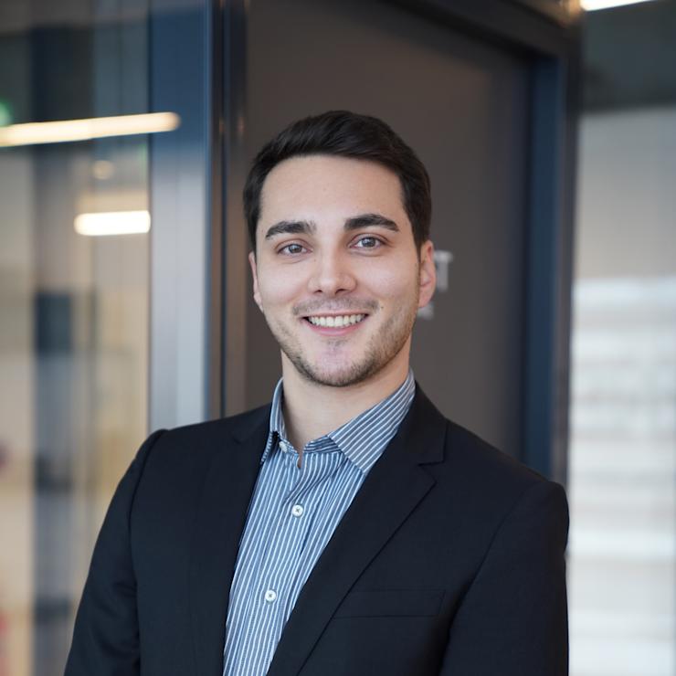 Ein junger Mann im Anzug steht lächelnd in einem modernen Innenraum.