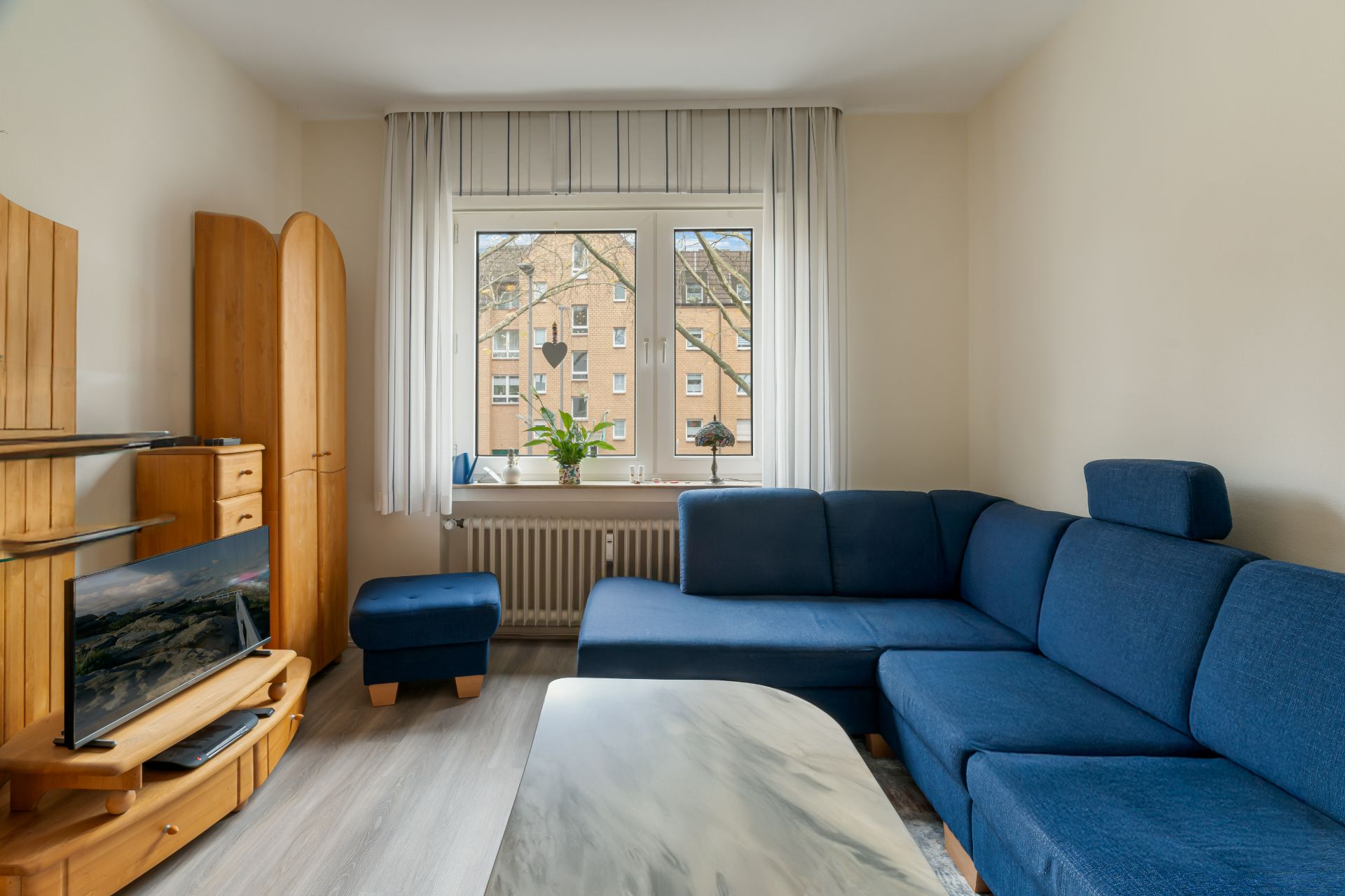 Wohnzimmer mit blauer Eckcouch, TV, Holzmöbeln und Fenster mit Blick auf Mietshaus.